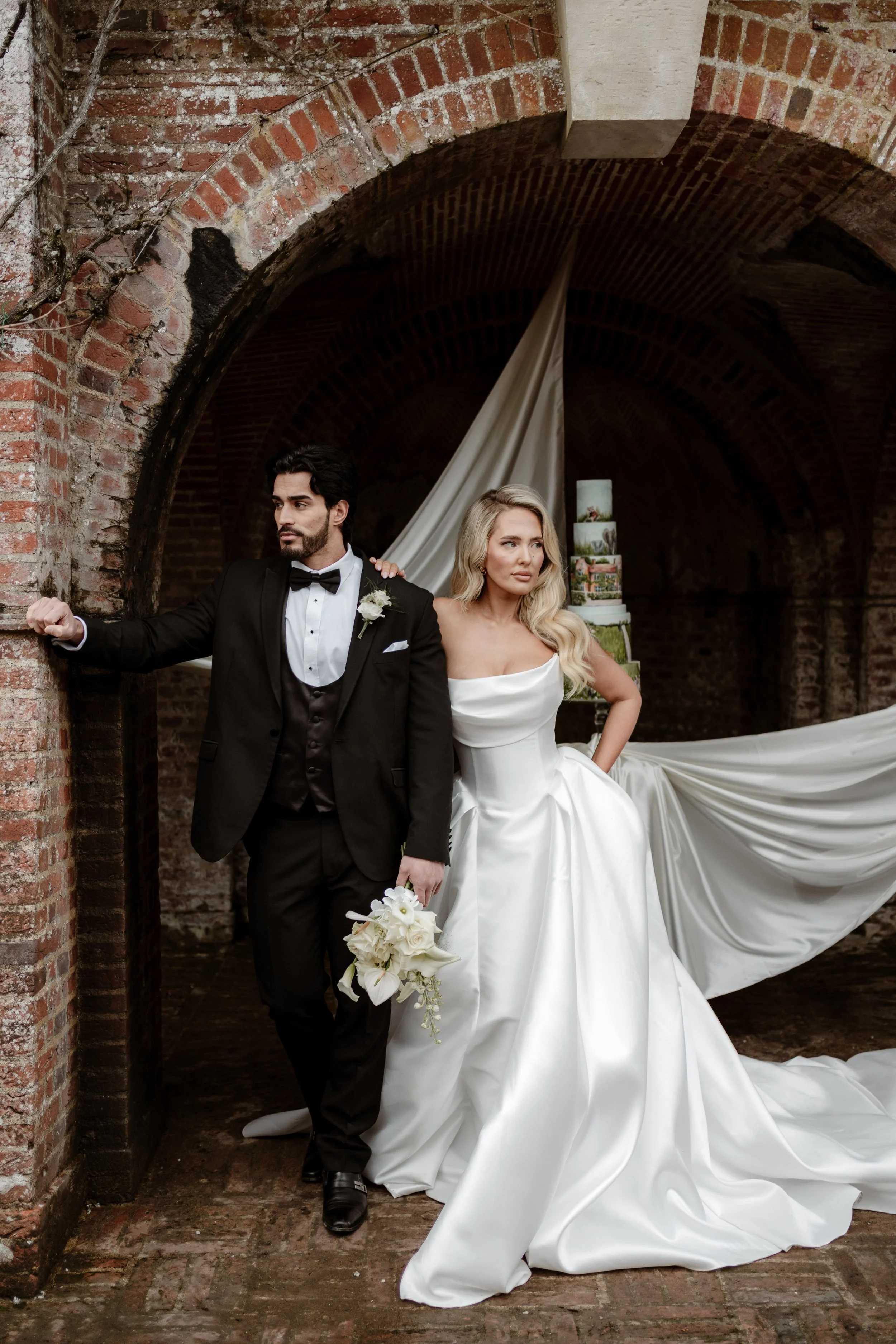 A bride and groom standing in an arched brick tunnel. The groom is in a black tuxedo and bowtie, holding a flower bouquet. The bride wears a white wedding gown with a long train, holding a bouquet of white flowers. There is a wedding cake visible in 