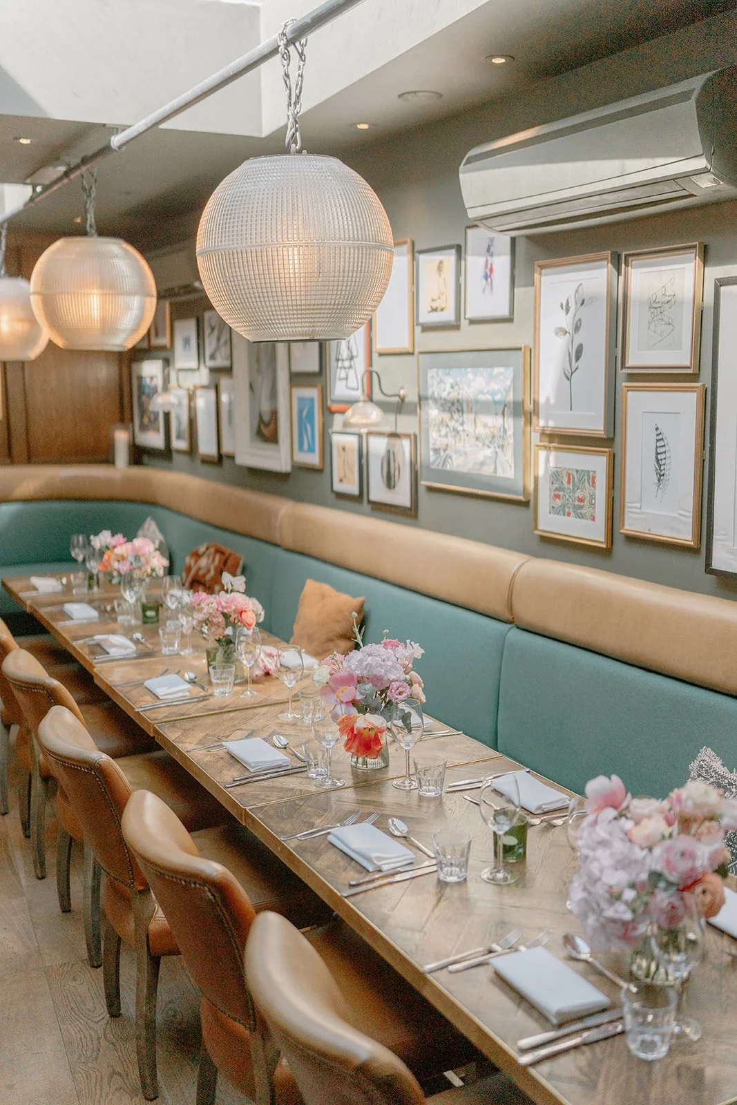 A long wooden dining table set for a meal with floral centerpieces, wine glasses, water glasses, napkins, and silverware in a decorated dining room with framed artwork and hanging globe pendant lights.