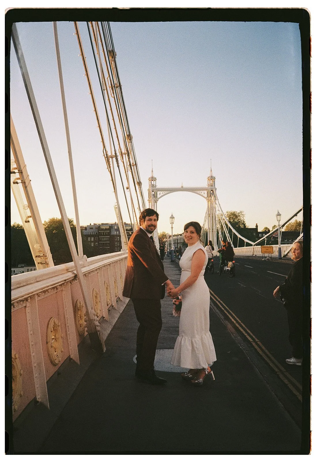 A couple holding hands on a bridge during sunset, with a cityscape and a large white suspension bridge in the background.