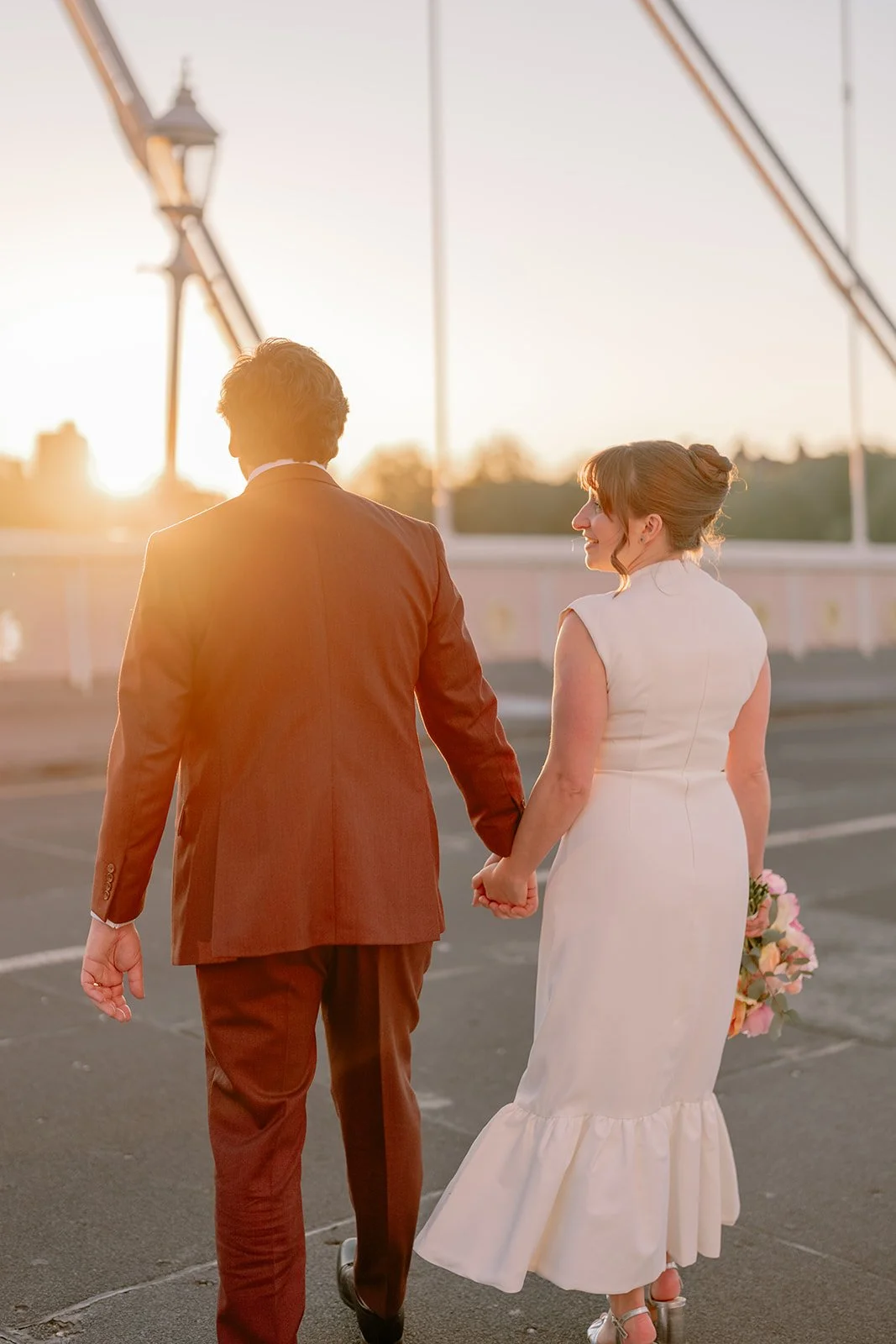 A newlywed couple holding hands and walking outdoors at sunset, one in a brown suit and the other in a white wedding dress, with a bridge in the background.