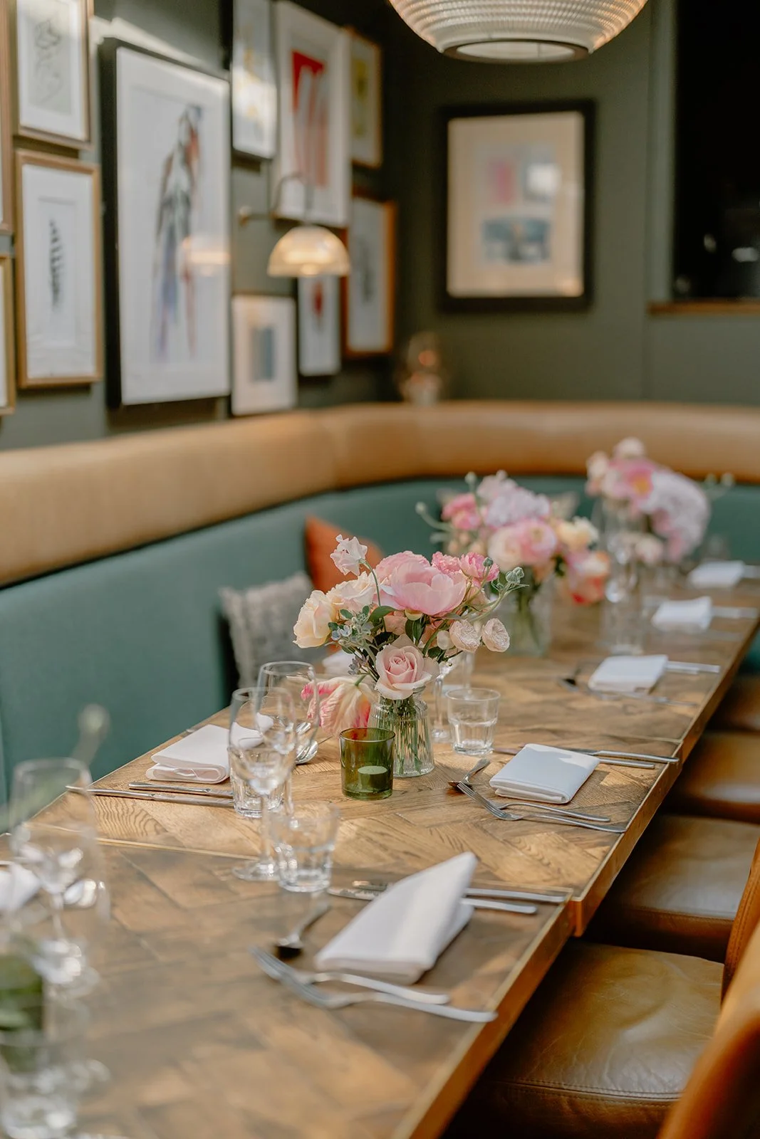 A restaurant table set with white napkins, wine glasses, water glasses, and silverware, decorated with pink and white flower arrangements in vases, with a green cushioned bench and framed art on a dark wall in the background.