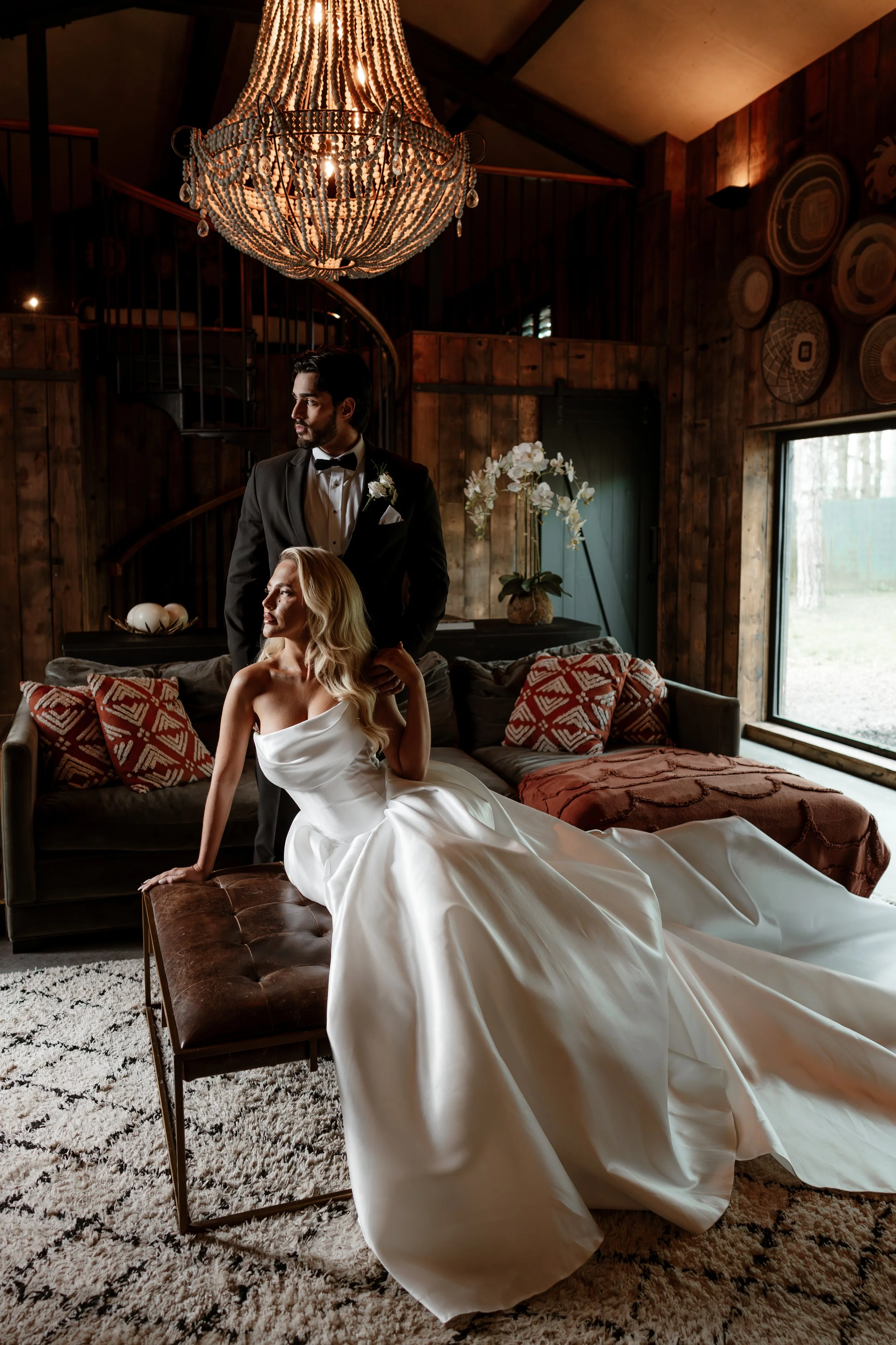 A bride in a strapless white wedding gown sitting on a bench with her hand on the floor, and a groom in a black tuxedo with a bow tie and white boutonniere standing behind her in a rustic wooden room with a large beaded chandelier, potted white orchi