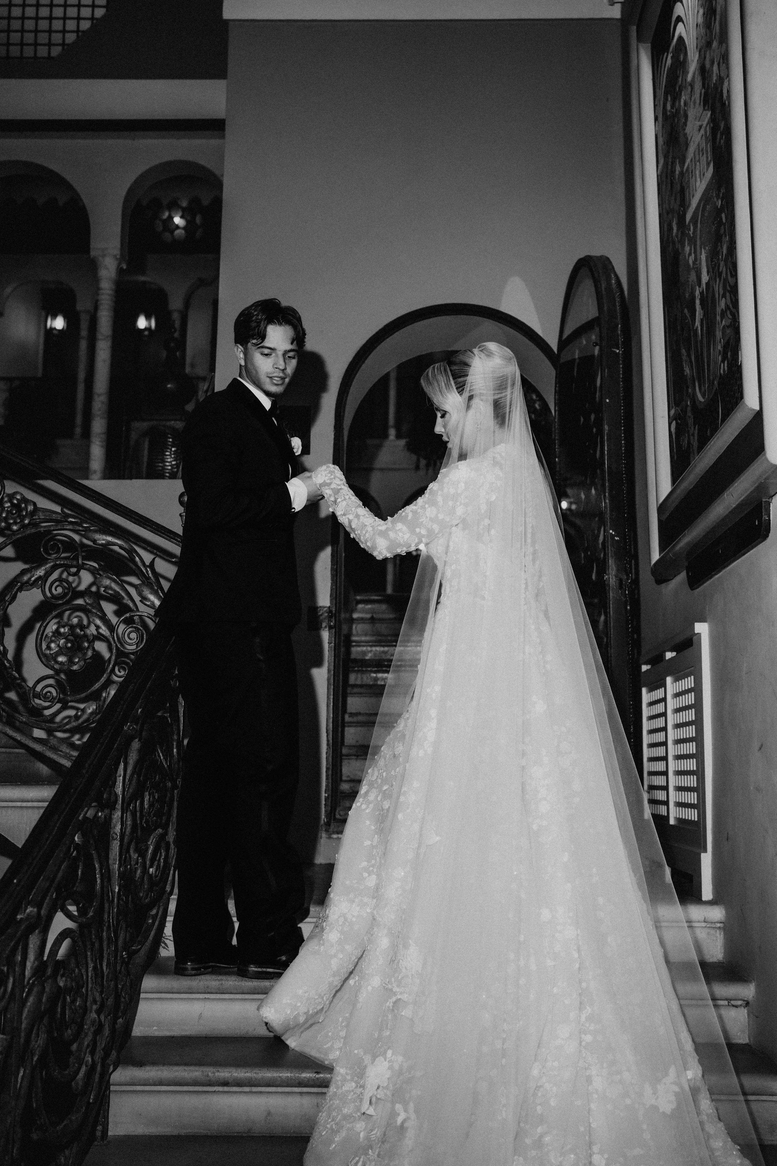 A black and white photo of a bride and groom holding hands on a staircase inside a building with ornate railings and arched doorways.