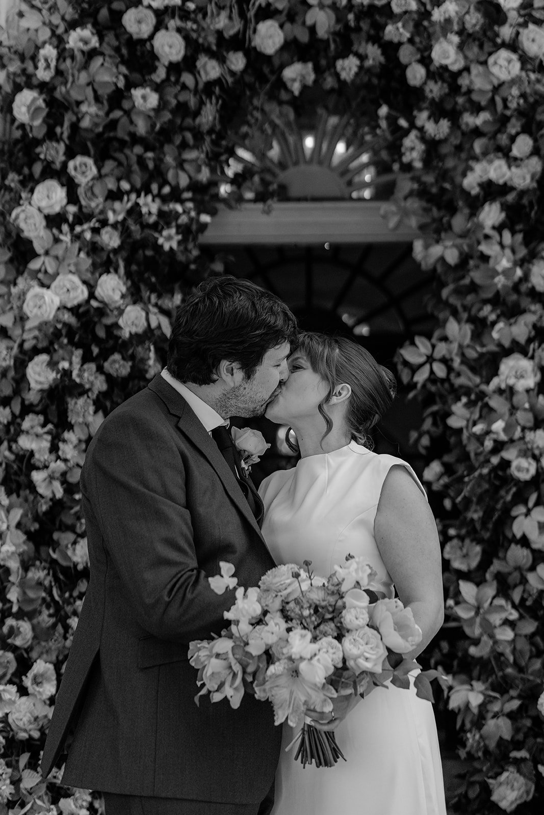 A black and white photo of a bride and groom kissing, with the bride holding a bouquet of flowers. They are standing in front of a floral arch.