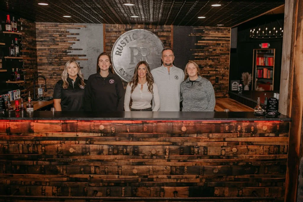 Group of five people standing behind a wooden bar in a rustic-themed establishment, with shelves of bottles on the left and a large metal sign behind them.