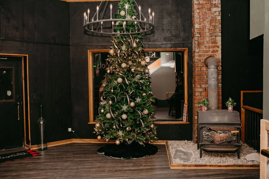 Decorated Christmas tree with gold and silver ornaments and ribbon in a cozy room with dark walls, a black chandelier, and a wood-burning stove.