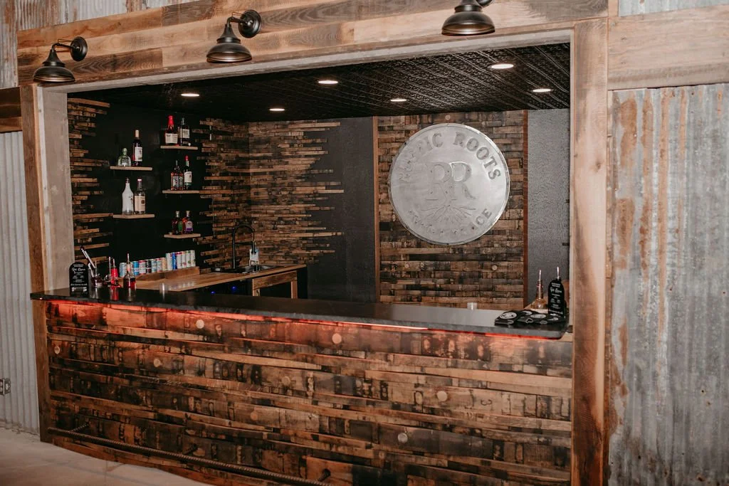 A rustic bar with a wooden front and back wall, shelves with liquor bottles, and a sign that reads "TRACTION ROOTS DR".