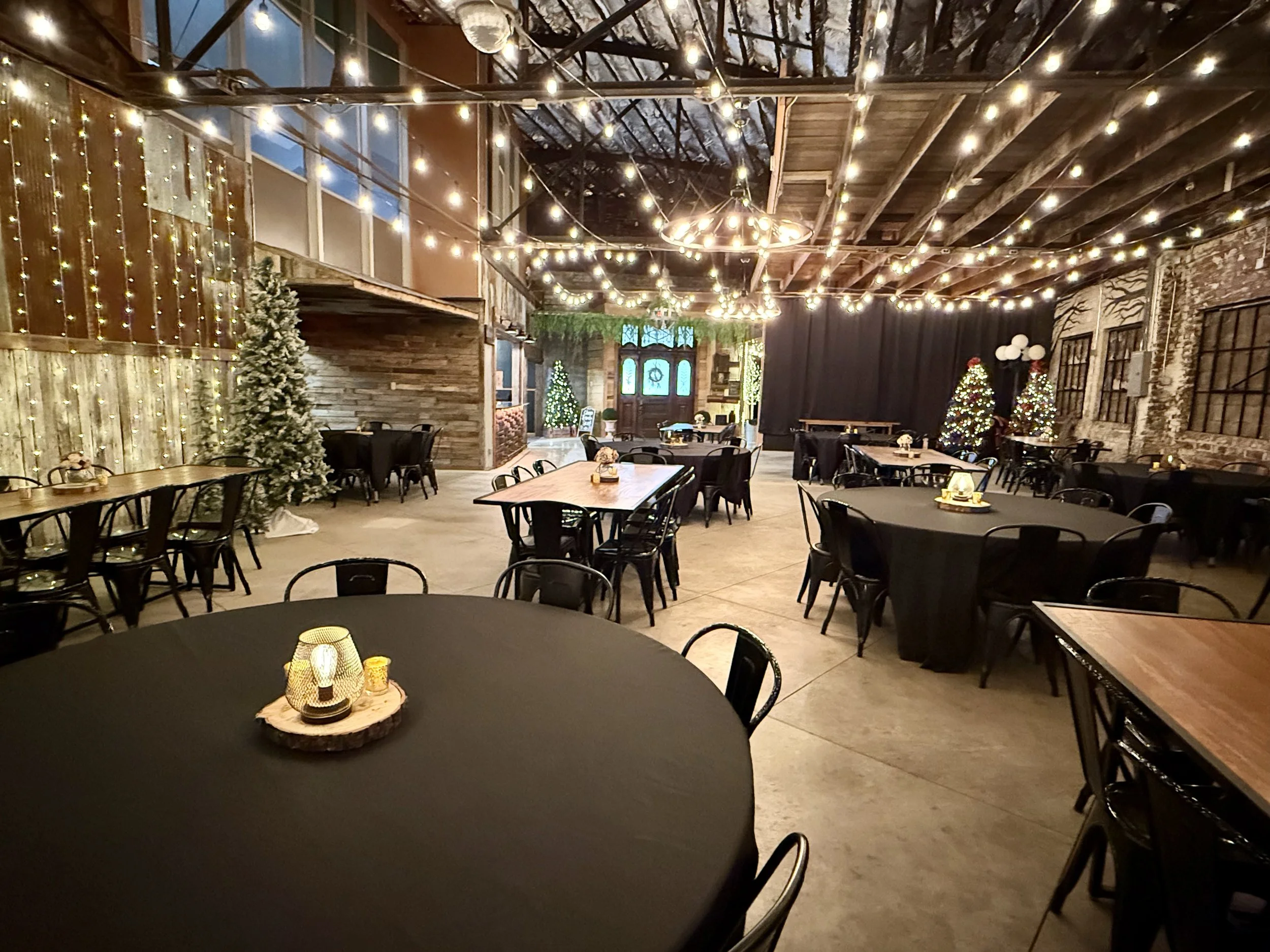Decorated event hall with string lights, Christmas trees, and festive candles on tables, ready for a celebration.
