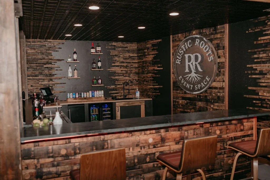 Rustic Roots event space bar area with wooden walls and bar, shelves with bottles, seating stools, and a large circular sign that says Rustic Roots Event Space.