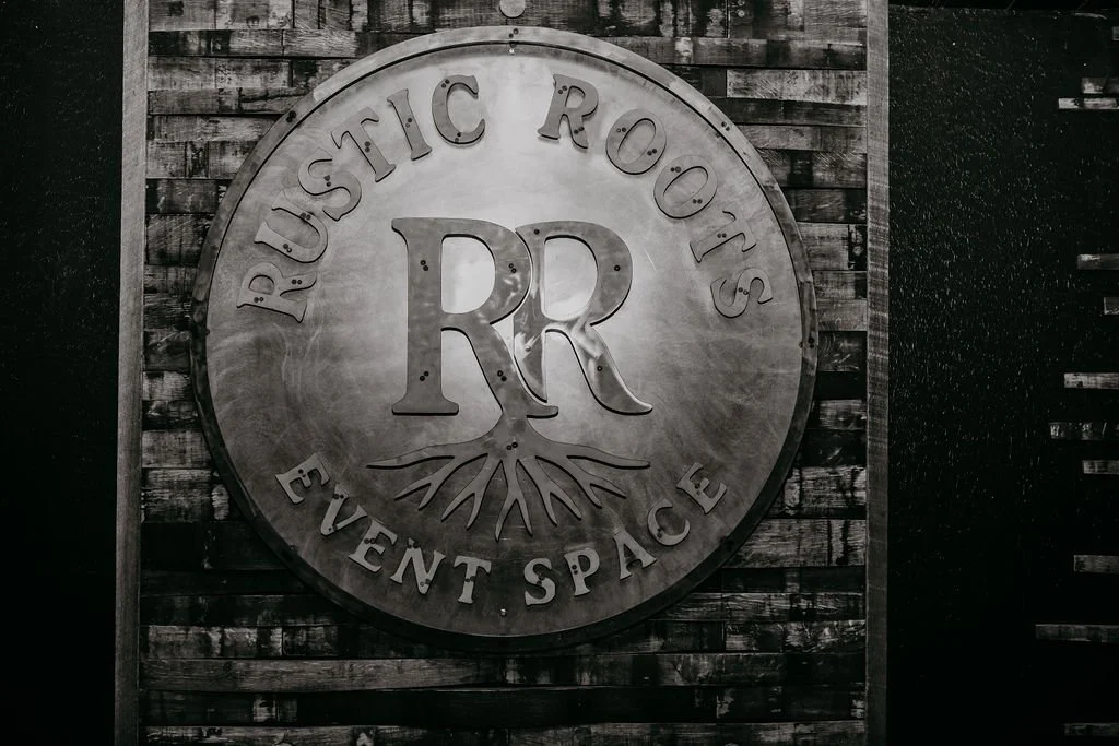 A large circular metal sign on a wooden wall displaying the text 'DUSTIC ROOTS EVENT SPACE' with a logo of a tree with roots, and the initials 'RR' in the center.
