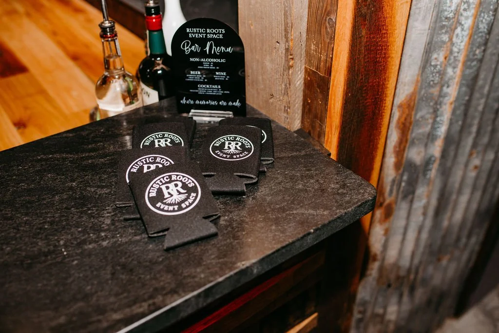 A bar area with a black coaster set, a small black sign listing drinks and non-alcoholic options, and bottles of liquor on a wooden surface.