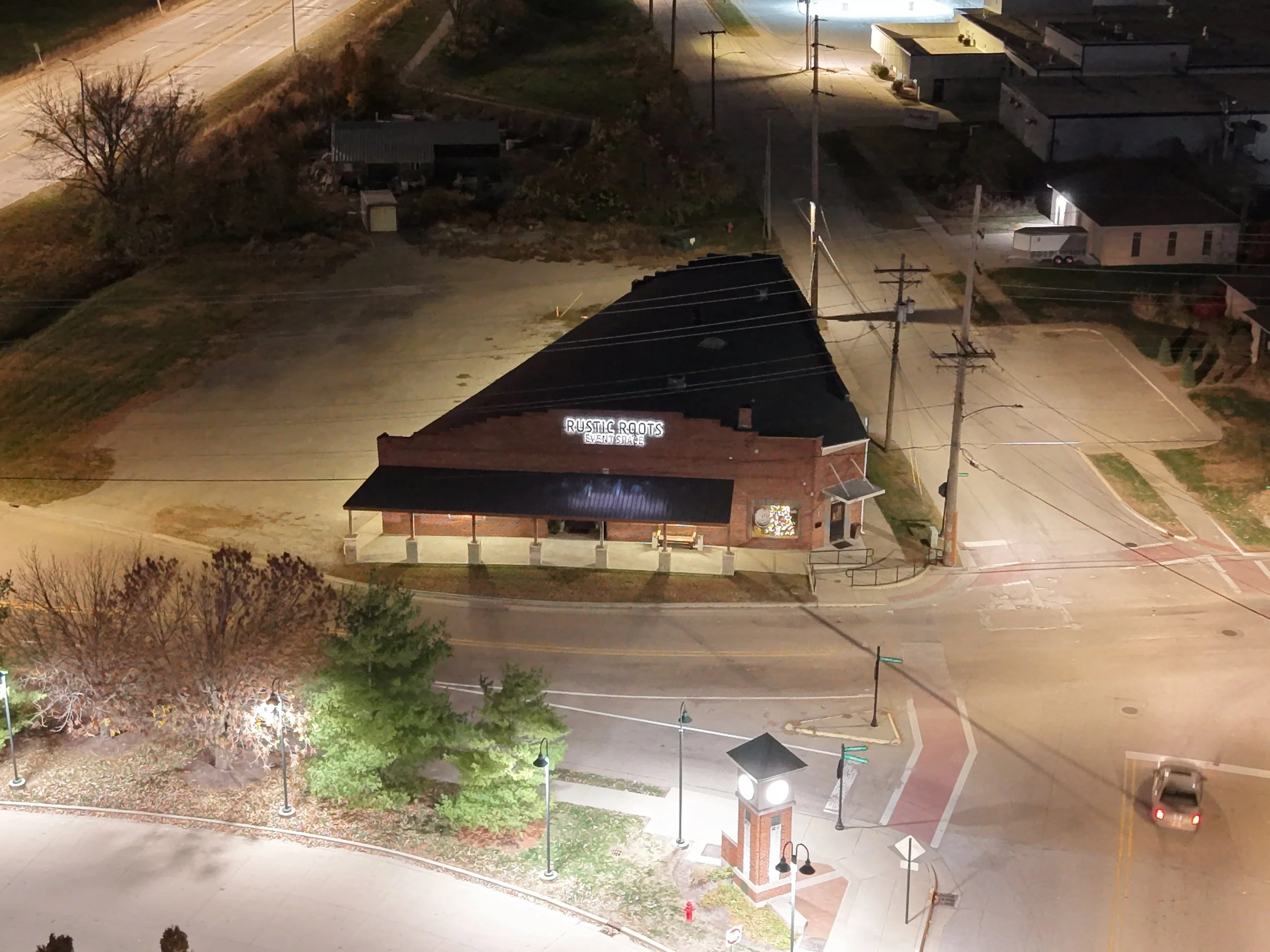 Night aerial view of a small brick building with a porch labeled 'Rustic Roots Event Space,' surrounded by streetlights, trees, parking lots, and a road with a car passing by.