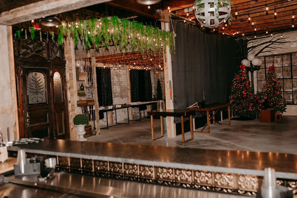 Festively decorated interior with Christmas trees, hanging string lights, and greenery, featuring a rustic design with wooden beams and a black curtain backdrop.