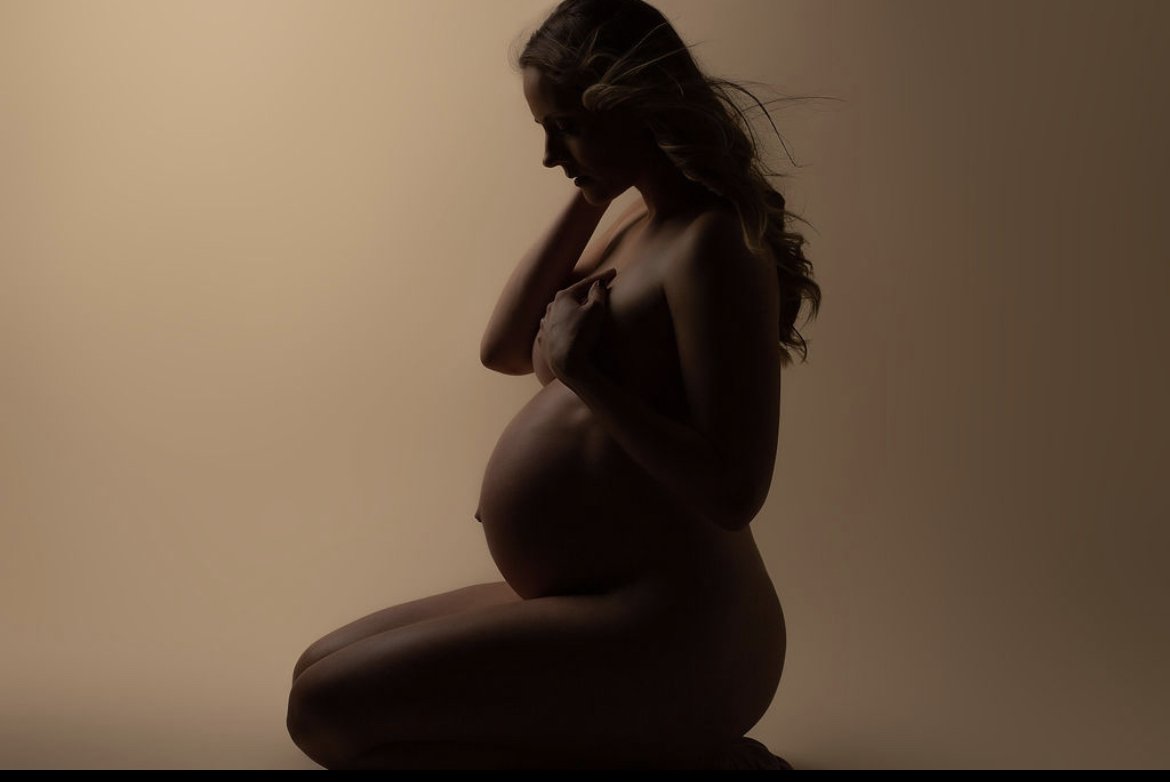 Silhouette of a pregnant woman kneeling, with her hands on her chest and hair flowing, against a plain background.