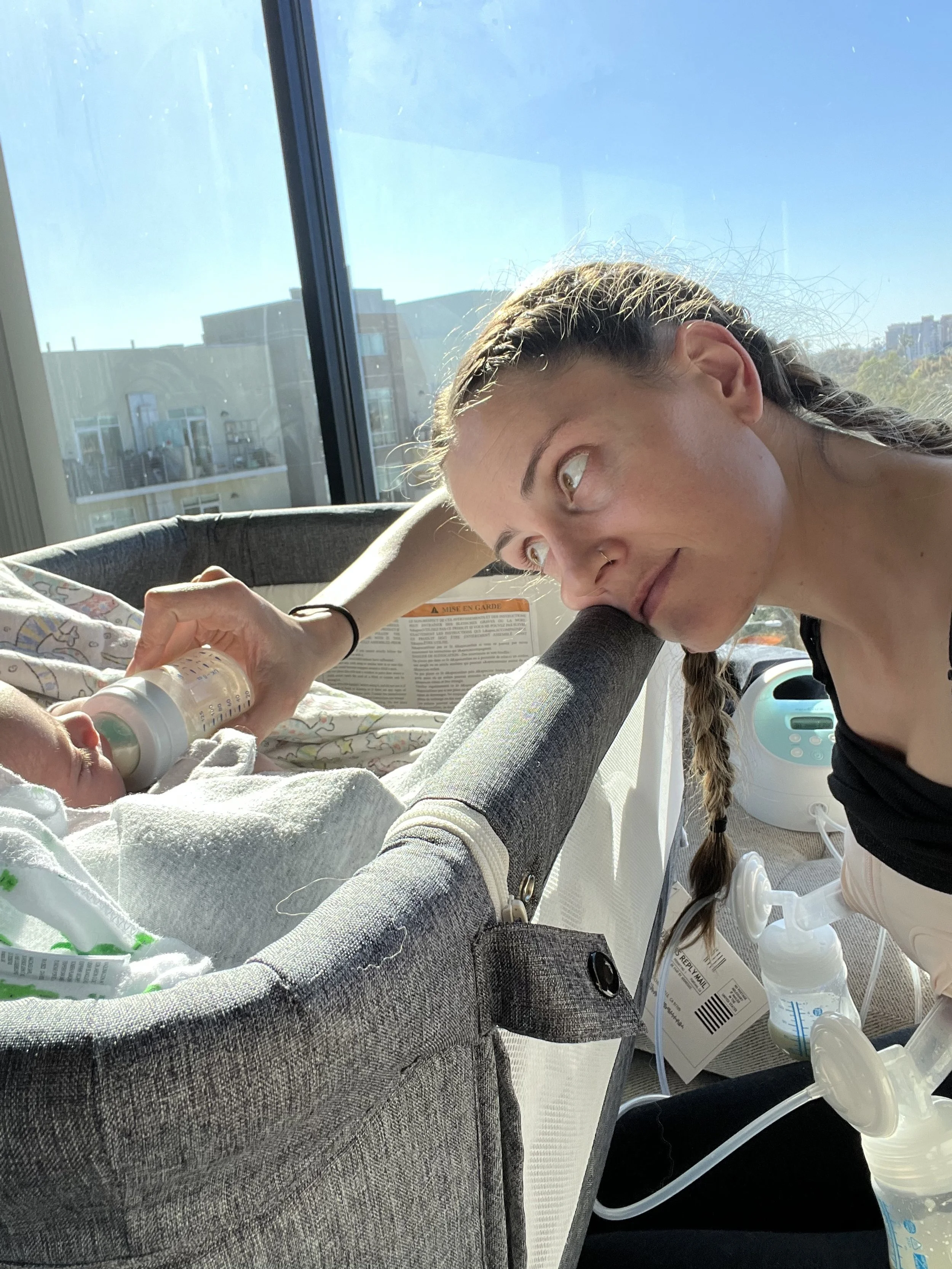 Young woman with a braid gently kisses a newborn baby in a hospital incubator, sunlight streaming through large windows behind them.