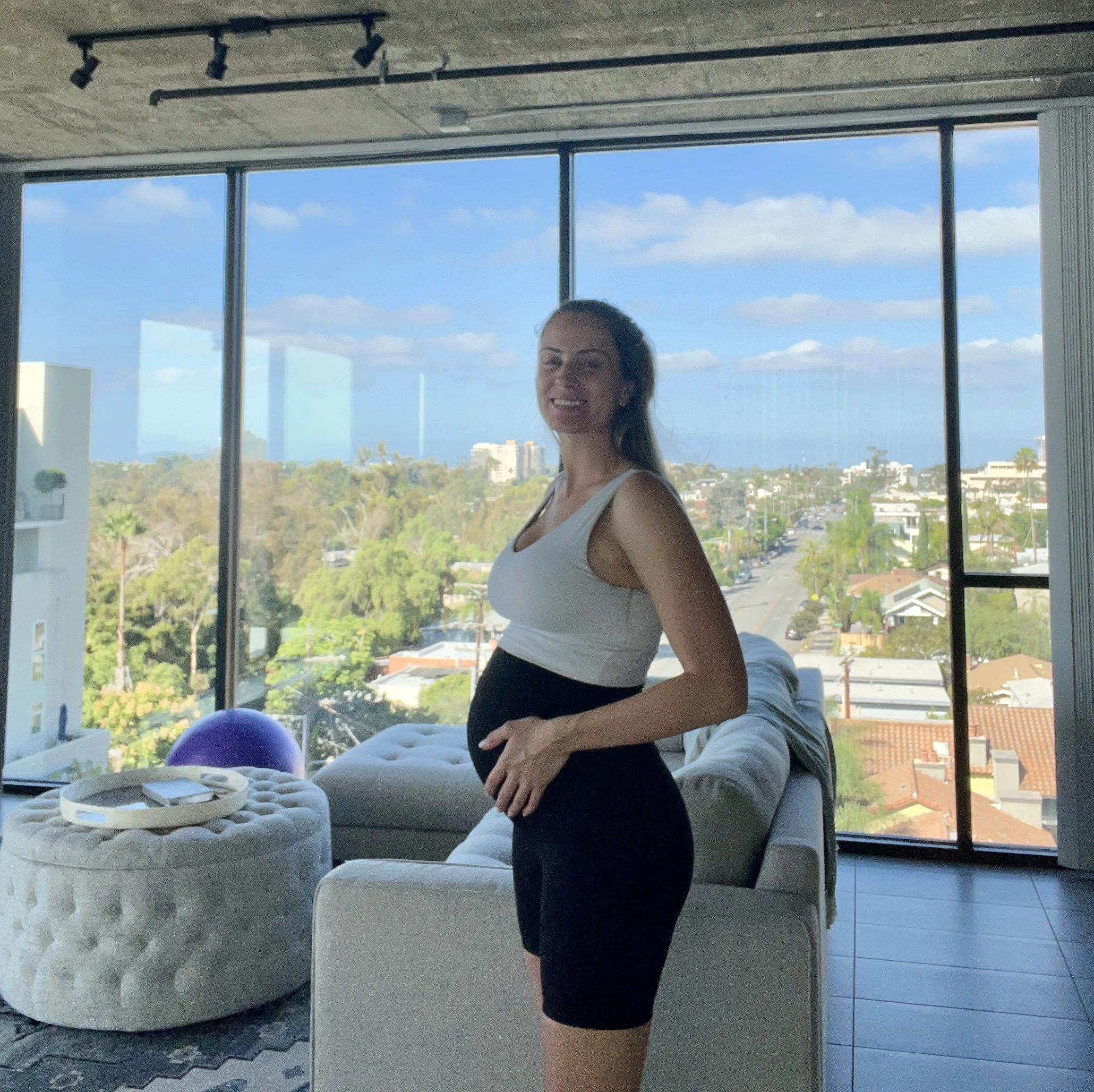 Pregnant woman in a white top and black shorts standing in a modern living room with large windows showing a cityscape and blue sky.