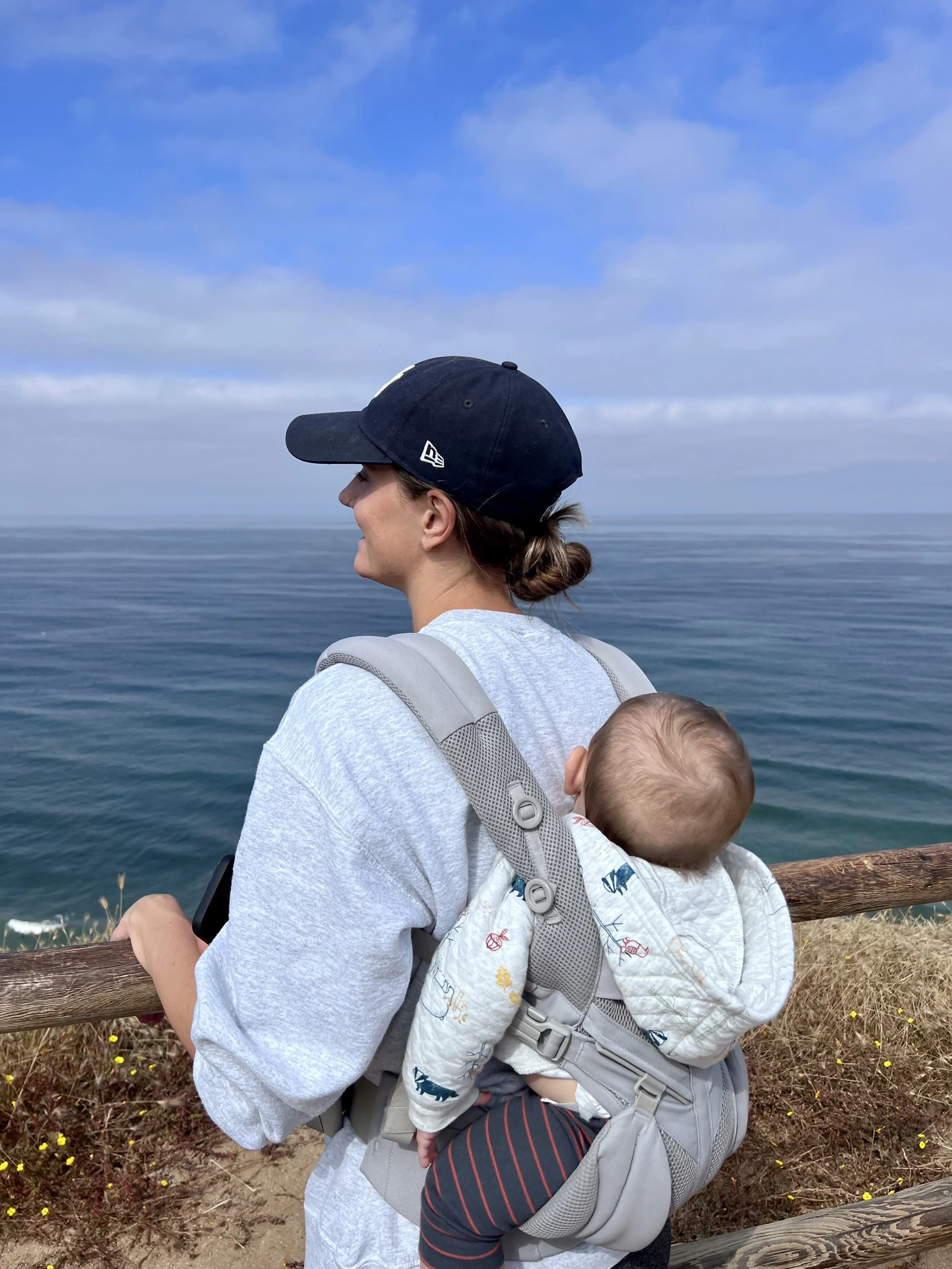 Woman with a baby in a backpack carrier looking out over the ocean from a wooden railing on a cloudy day.