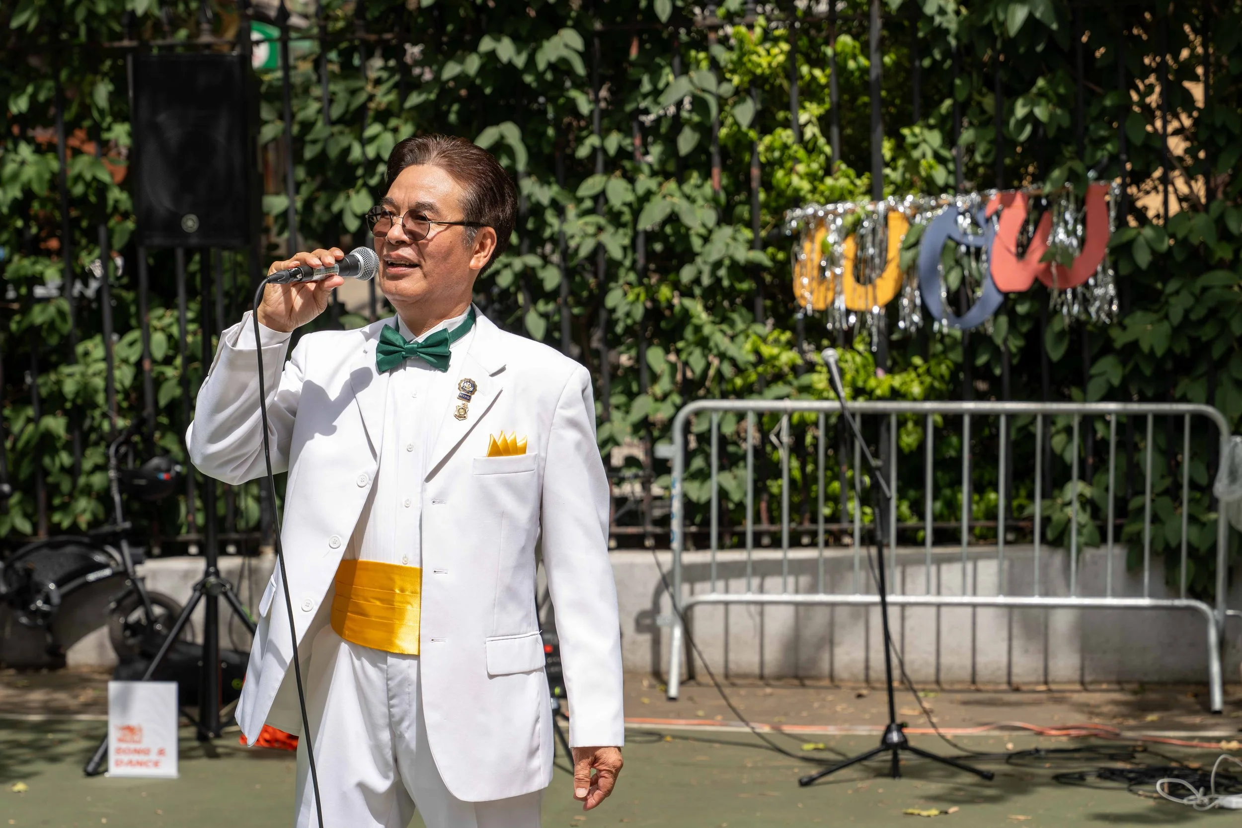 A man in a white tuxedo with a green bow tie and yellow sash, speaking into a microphone outdoors. Background includes a decorative banner, green foliage, and a metal barricade.