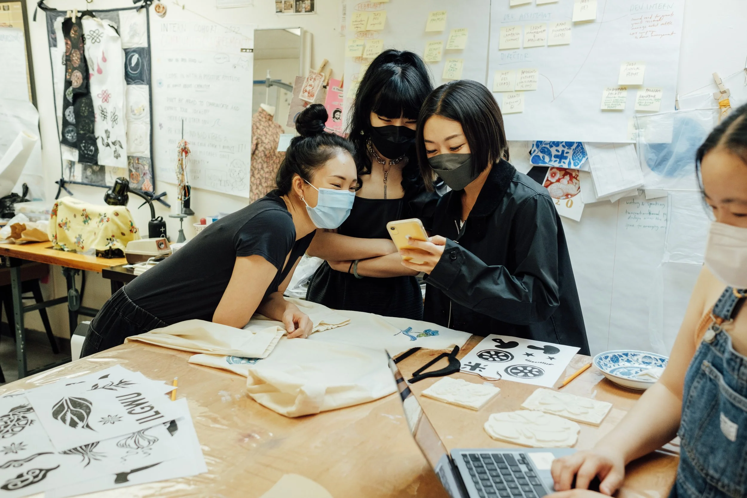 Four women wearing face masks gathered around a table, looking at a cell phone, in a creative workspace with design sketches and projects on the wall.