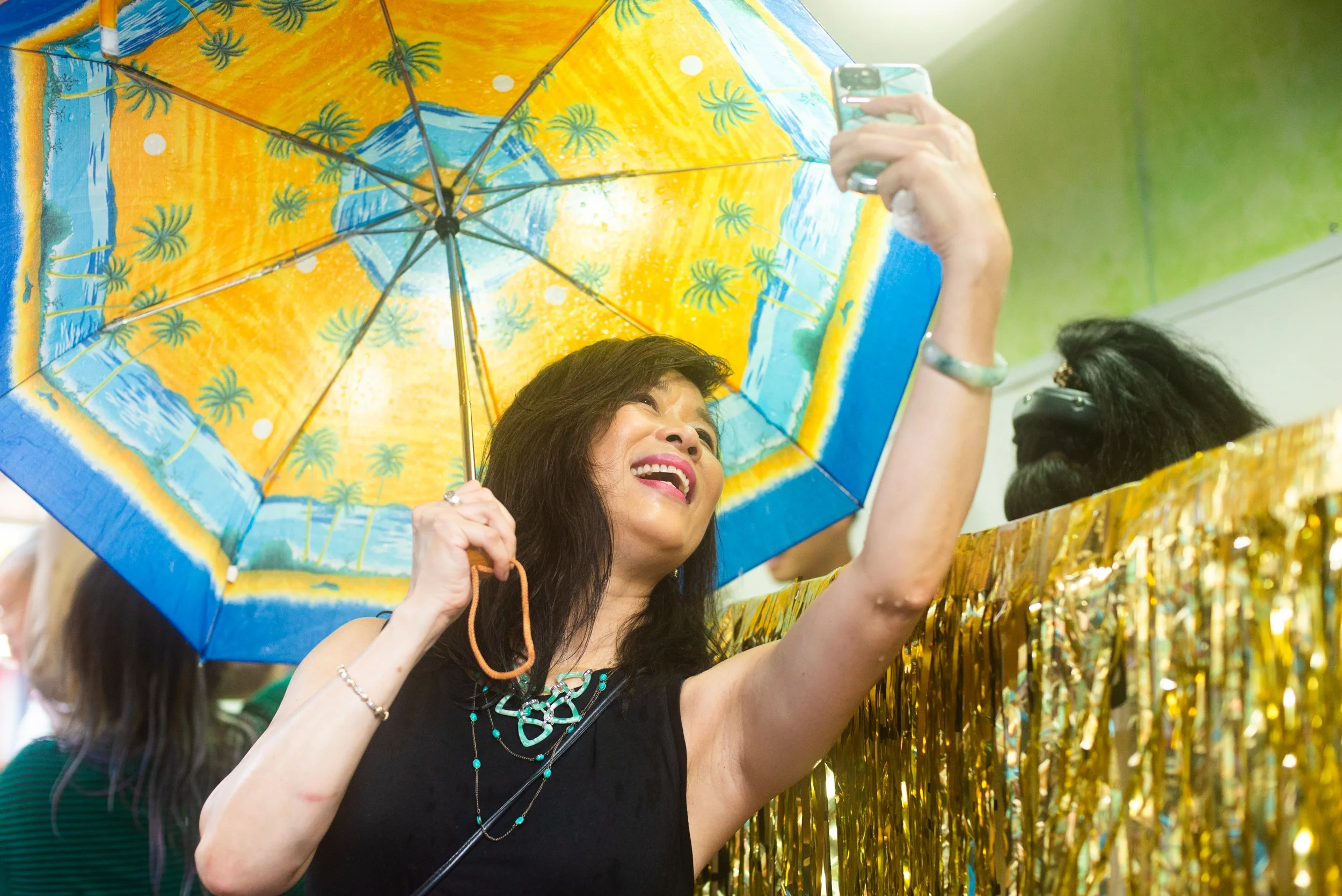 Woman smiling and taking a selfie with a colorful beach-themed umbrella, standing behind a gold tinsel curtain.