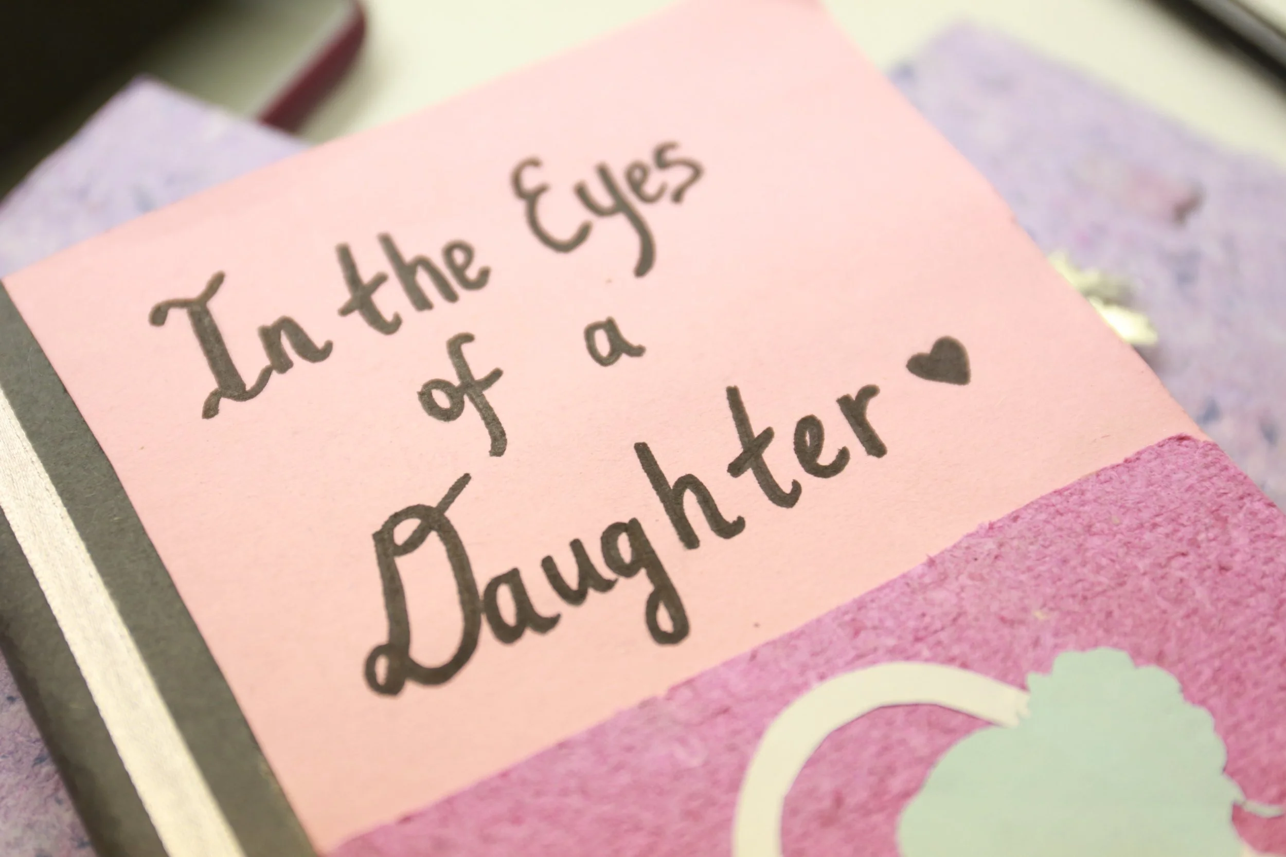 A pink card with handwritten text that reads 'In the Eyes of a Daughter' and a small heart symbol.