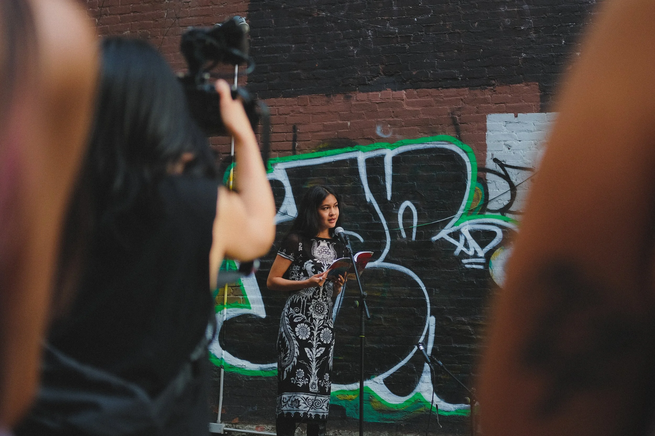 A woman in a black and white patterned dress reads from a book into a microphone on stage, with a graffiti-covered brick wall in the background. The photo is taken from between some audience members.