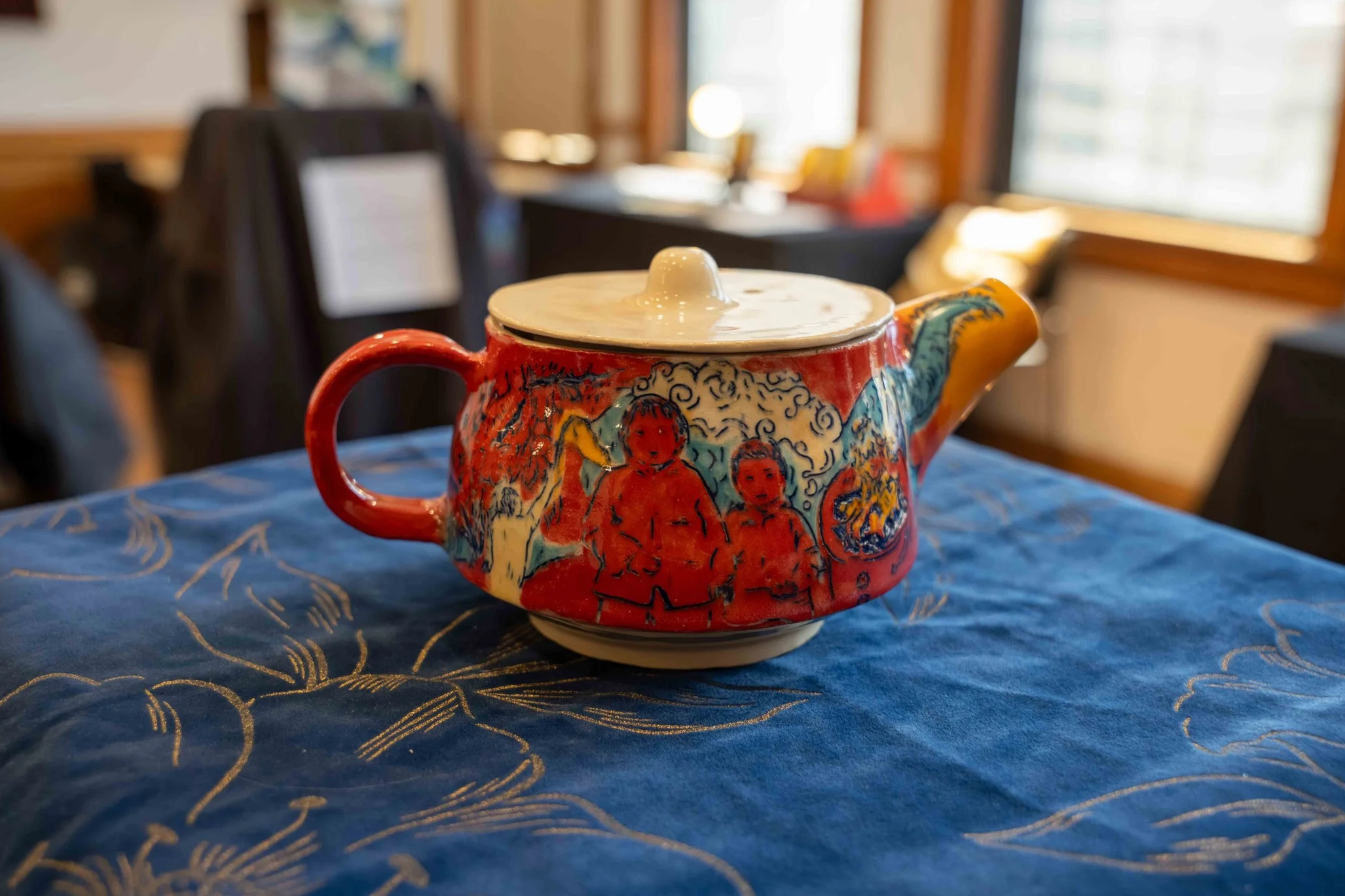Colorful ceramic teapot with drawings of children and vibrant artwork, placed on a blue patterned cloth in a cozy room with wooden walls and large windows.