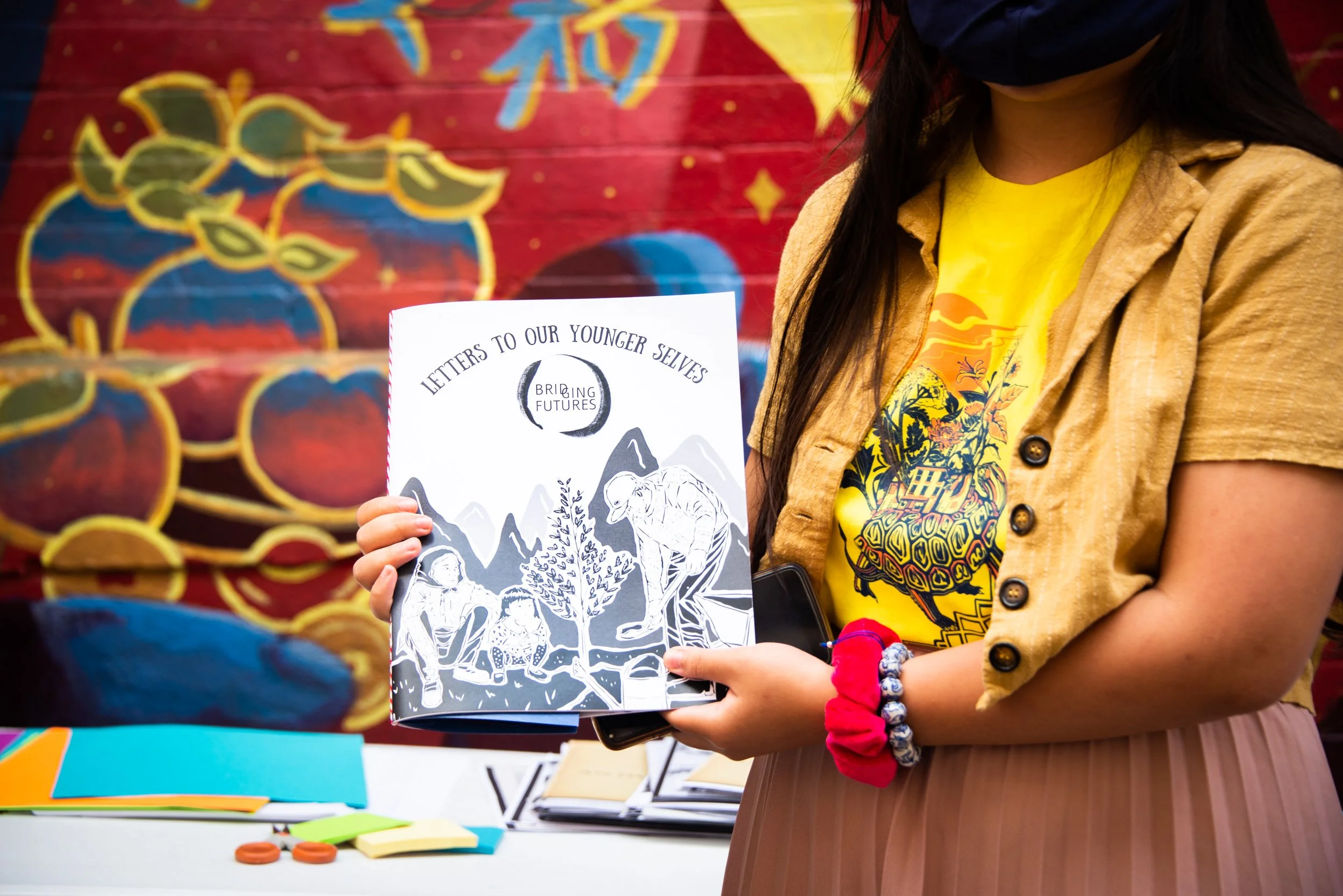 Person holding a booklet titled 'Letters to Our Younger Selves' with illustrations of children and mountains on the cover, standing in front of a colorful background with balloons.