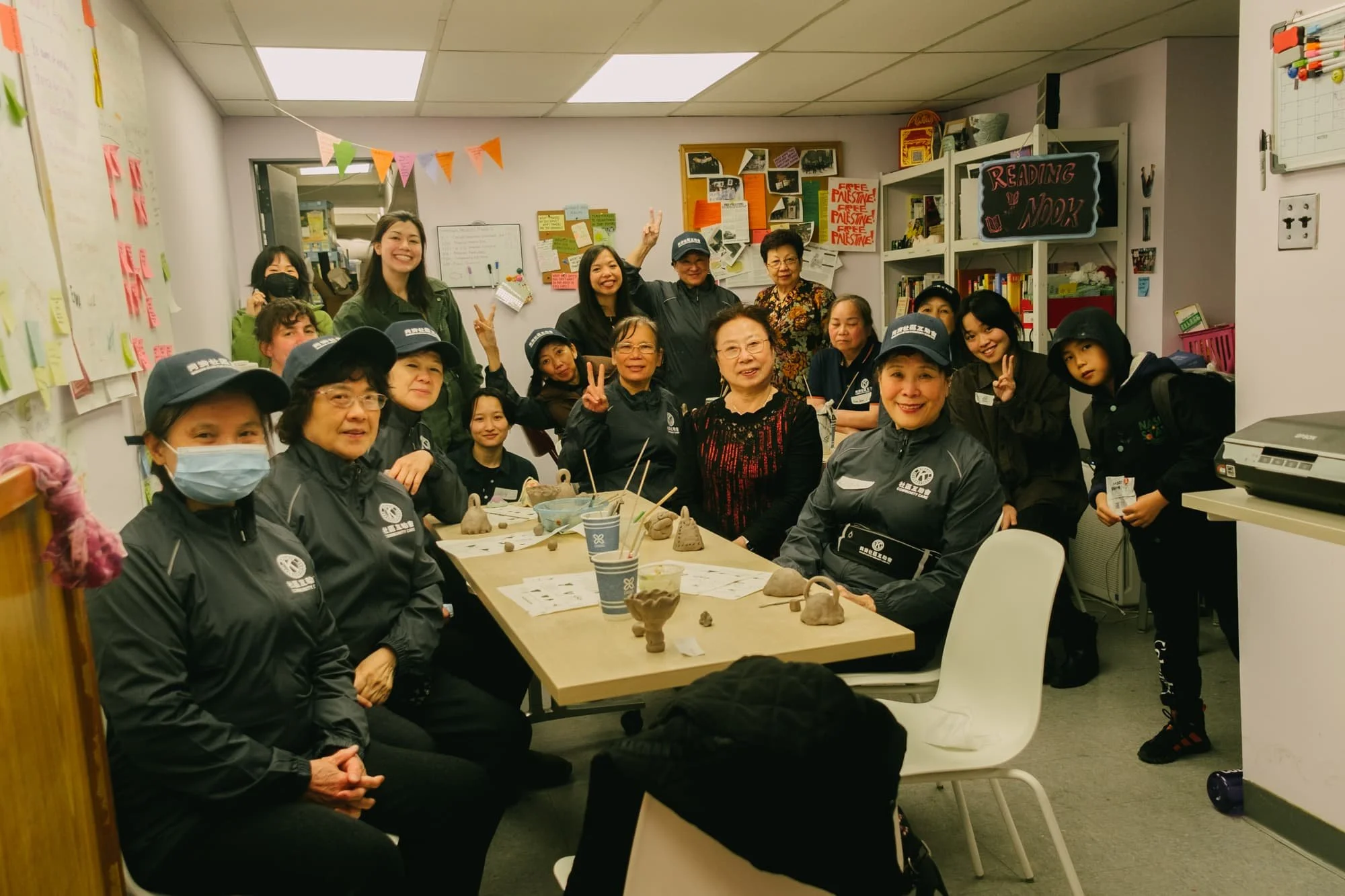 Group of women and girls gathered around a table in a classroom or community room, some wearing matching gray jackets and caps with logos, with some making peace signs, and others smiling at the camera. The table has clay projects and cups, and the room has colorful posters and a bookshelf with a sign that says 'Reading Nook'.