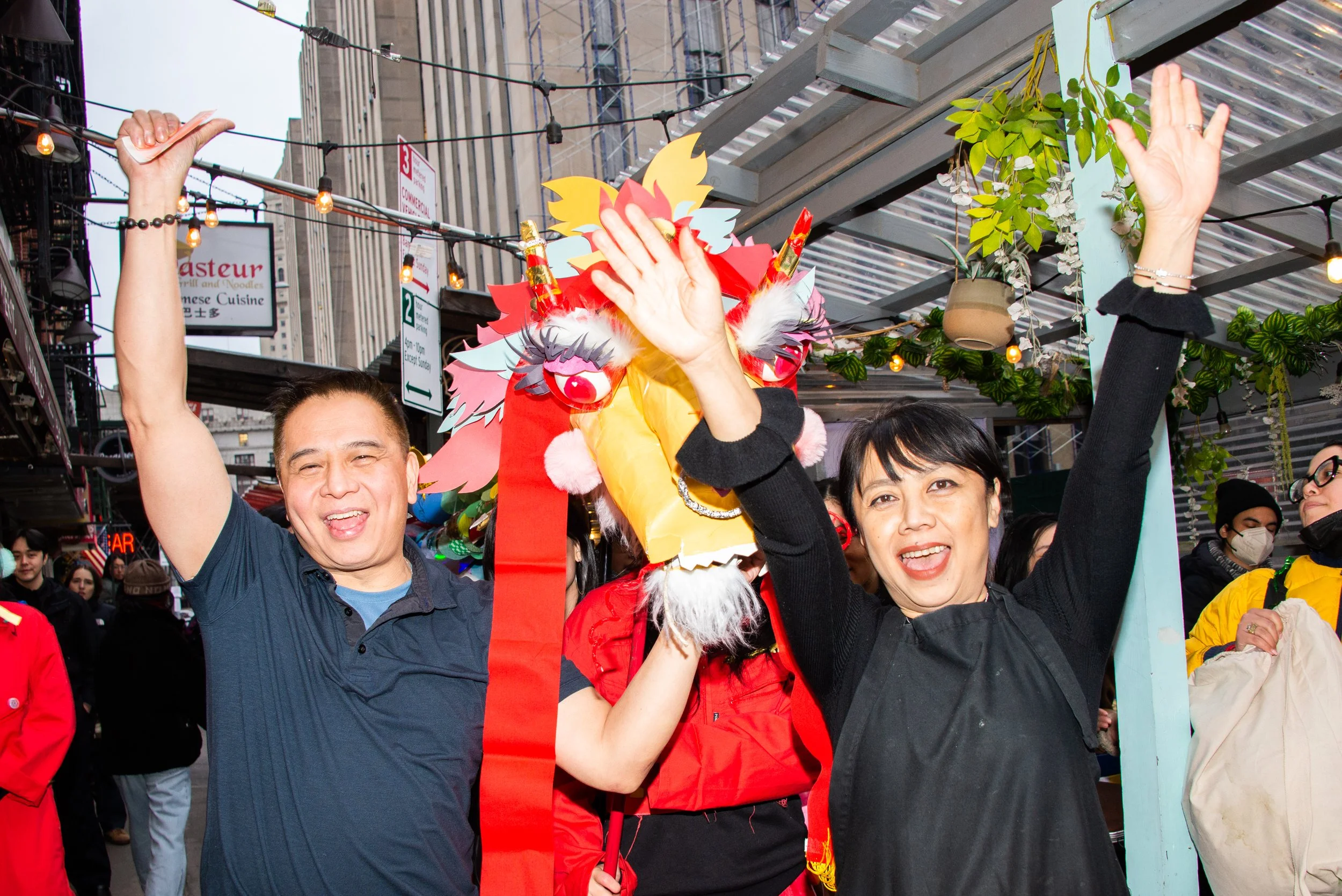 Two people celebrating during a cultural event with a colorful dragon costume in an urban setting, surrounded by a crowd, string lights, and city buildings.