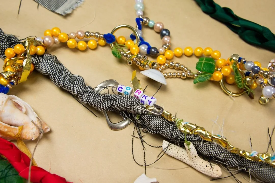 Collection of colorful beaded necklaces and jewelry on a beige surface, including a black and white braided strap, gold accents, and small decorative elements.