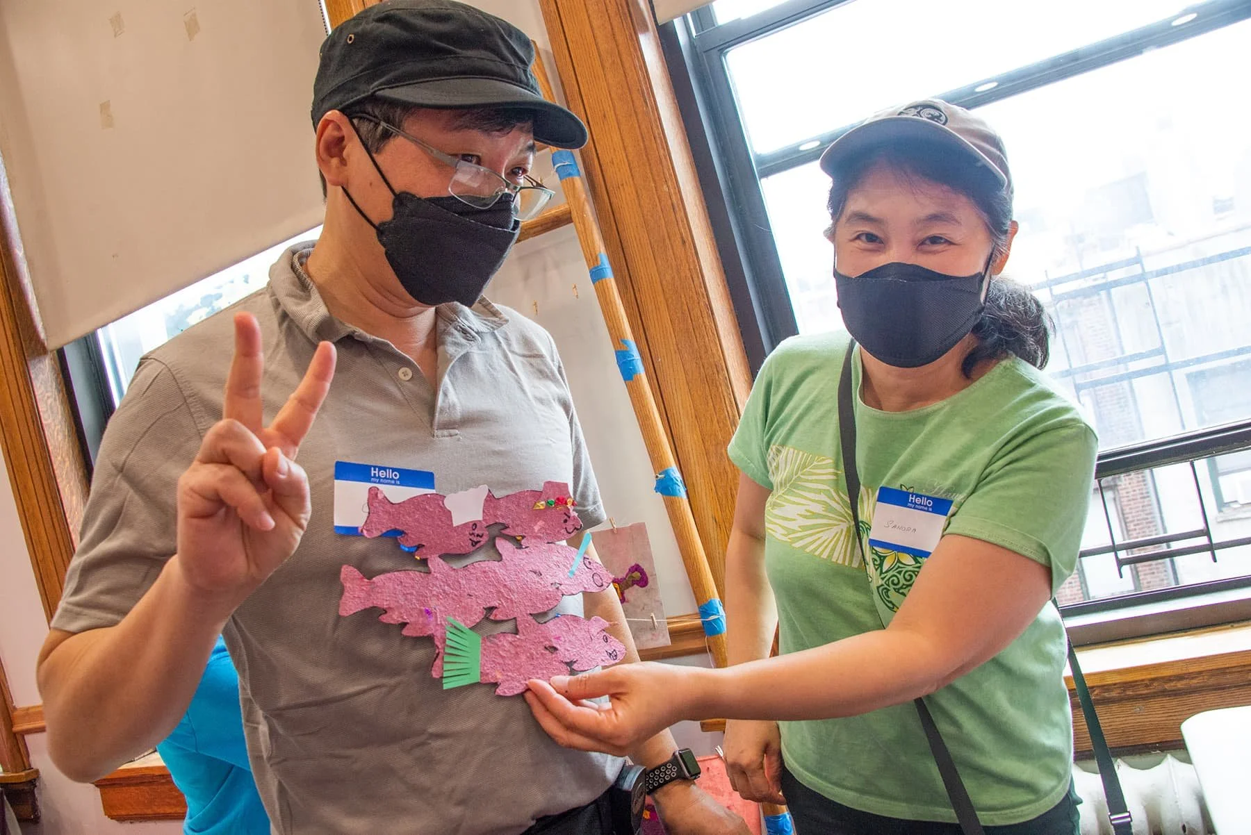 Two women wearing masks, standing indoors near a window and holding a pink paper craft resembling a group of animals with colorful decorations, one making a peace sign.