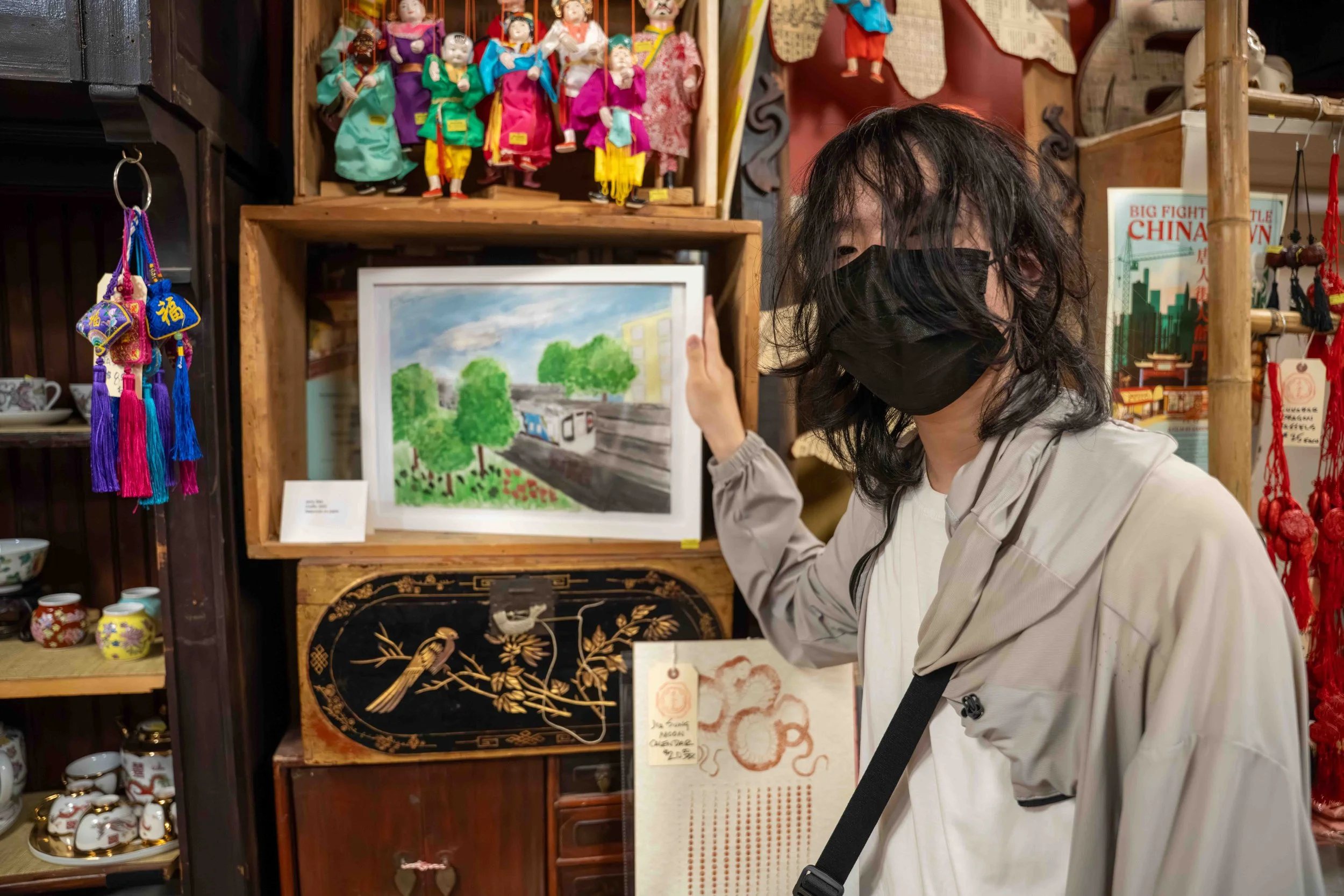 Person with black hair wearing black face mask and light-colored jacket pointing at framed watercolor painting of a train and greenery in a store with Asian decor and souvenirs.
