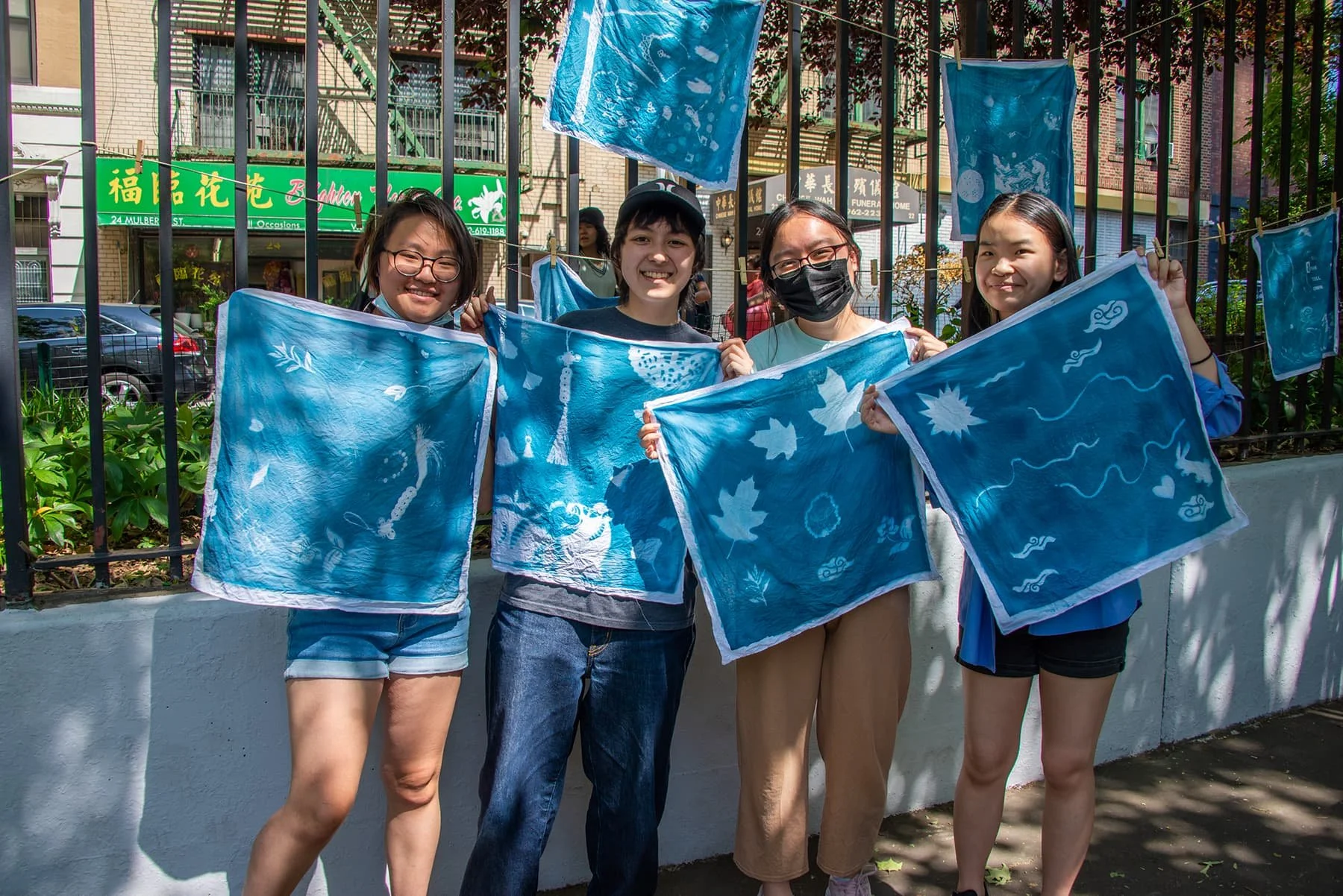 Four young women smiling and holding blue fabric with white designs outdoors in front of a black metal fence with hanging blue fabric. Behind them is a green sign with Chinese characters and a parking lot.