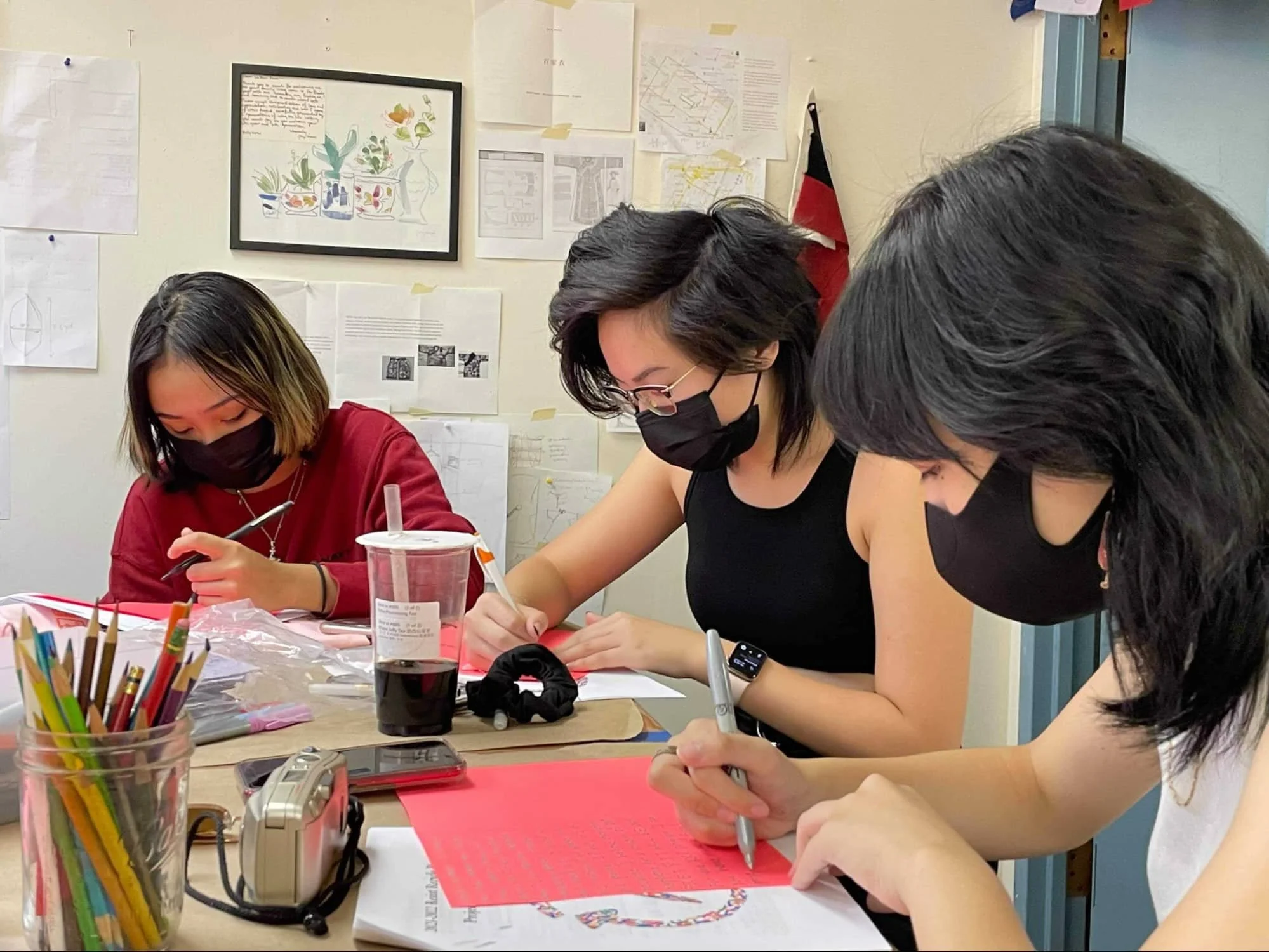 Three young women wearing black masks are sitting at a table, engaged in writing and drawing on paper, with art supplies and a drink on the table, in a room with various papers and drawings on the wall.