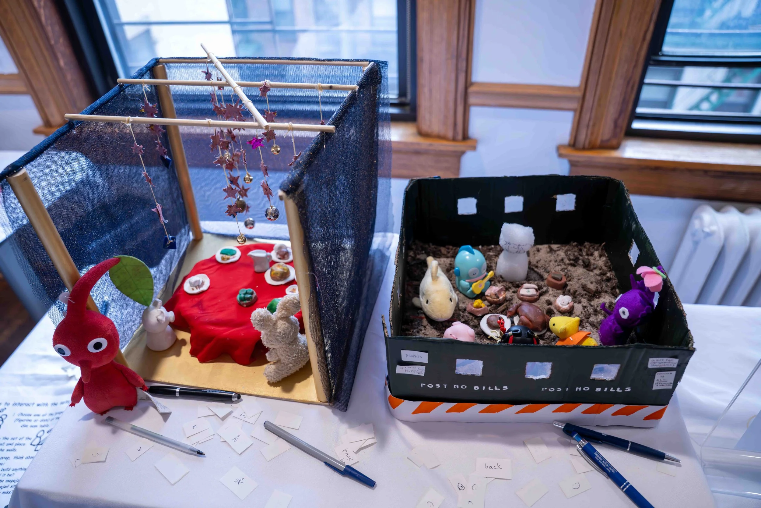 Two dioramas on a table. The left diorama depicts a miniature scene with a red cloth, small toys, and hanging decorations. The right diorama shows a miniature outdoor scene with small toys and rocks. Pens and sticky notes are scattered on the table in front.
