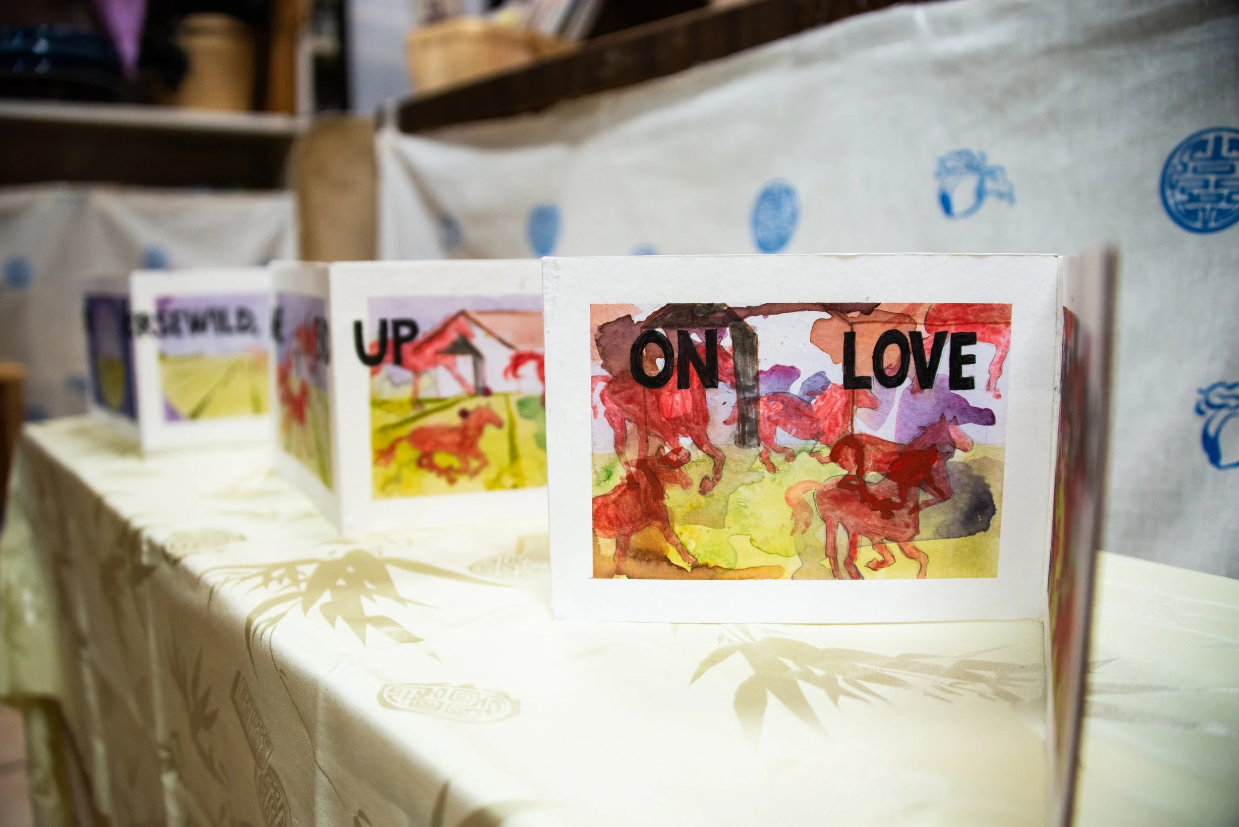 Display of colorful folded paper cards on a table, each featuring watercolor illustrations of horses and words like 'ON', 'LOVE', and 'UP' visible in the forefront.