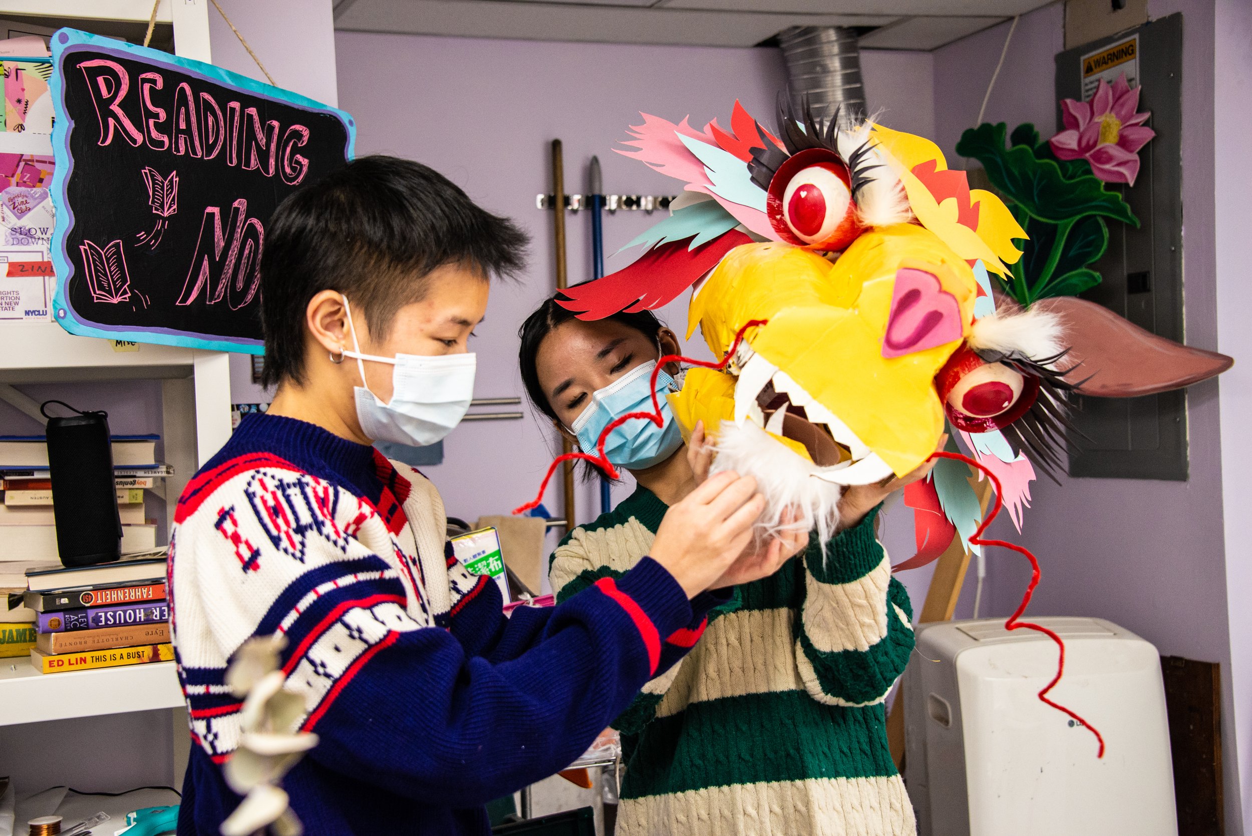 Two people wearing face masks are working on a colorful dragon puppet in a room with shelves of books and a sign that reads 'READING NO' in colorful chalk.