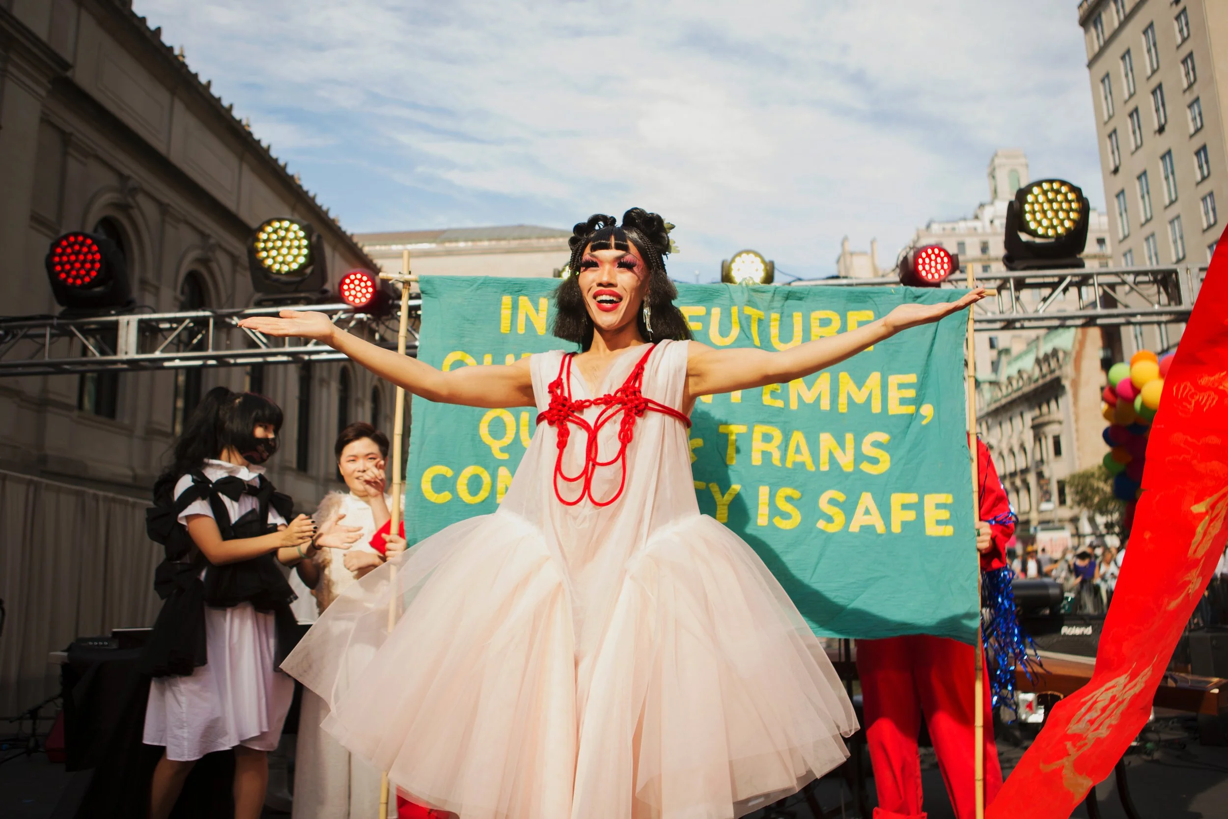 A person dressed in a flowing white dress with red accents, standing with arms outstretched on a stage during a celebration or protest at a city street, with a green banner and colorful balloons in the background.