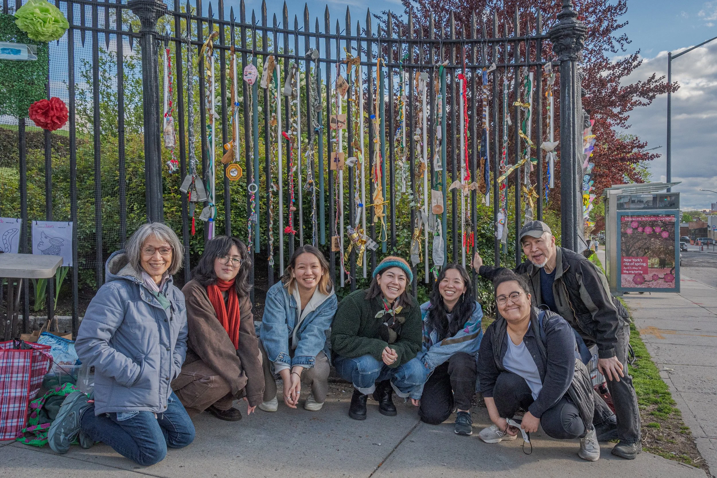 A group of seven people squatting and smiling in front of a black fence decorated with colorful ribbons and fabric pieces, outside on a sidewalk during daytime.
