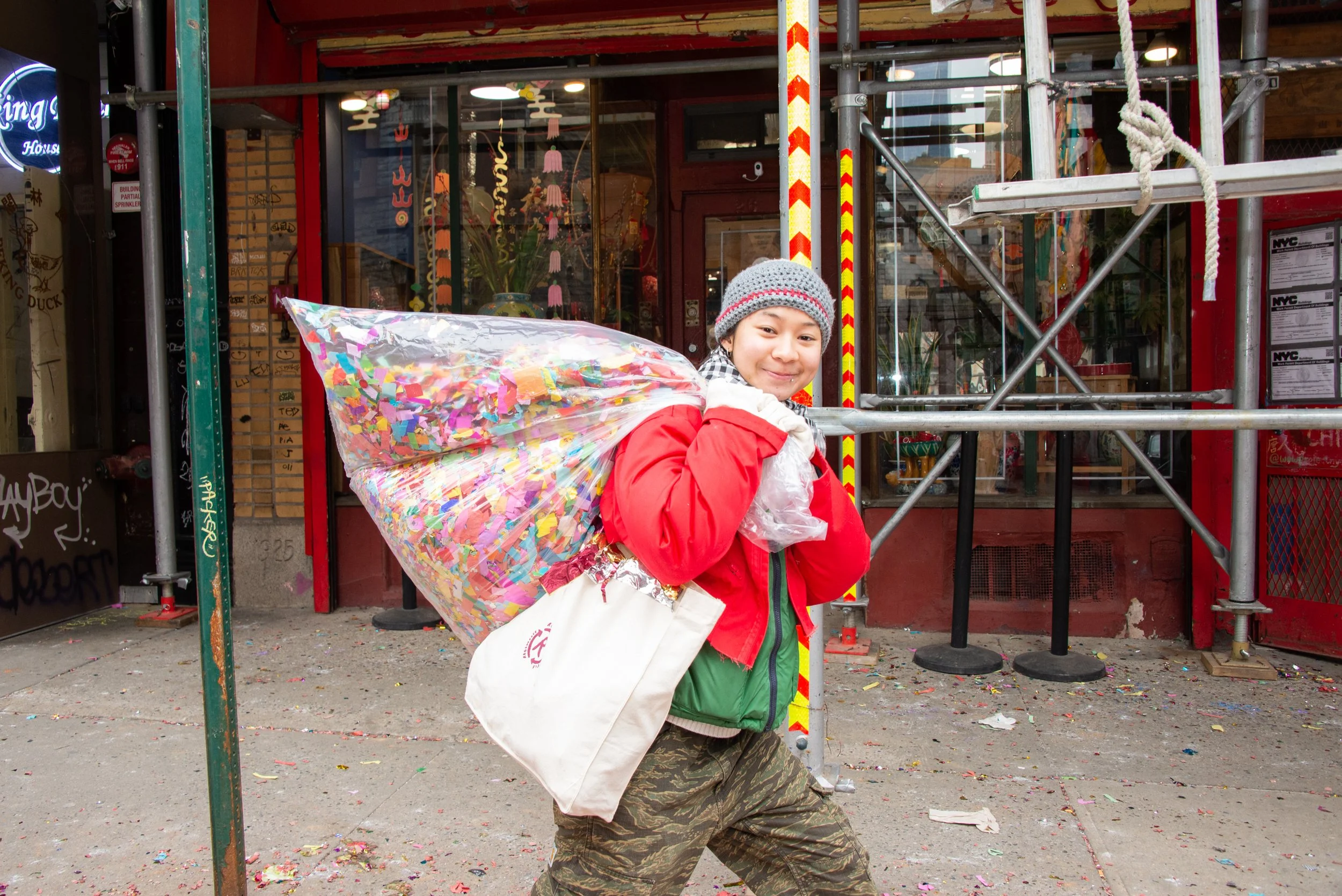 A young boy wearing a gray knit hat, a red jacket, and camouflage pants holding a large bag of colorful confetti on his shoulder, standing outside a store decorated for a celebration.