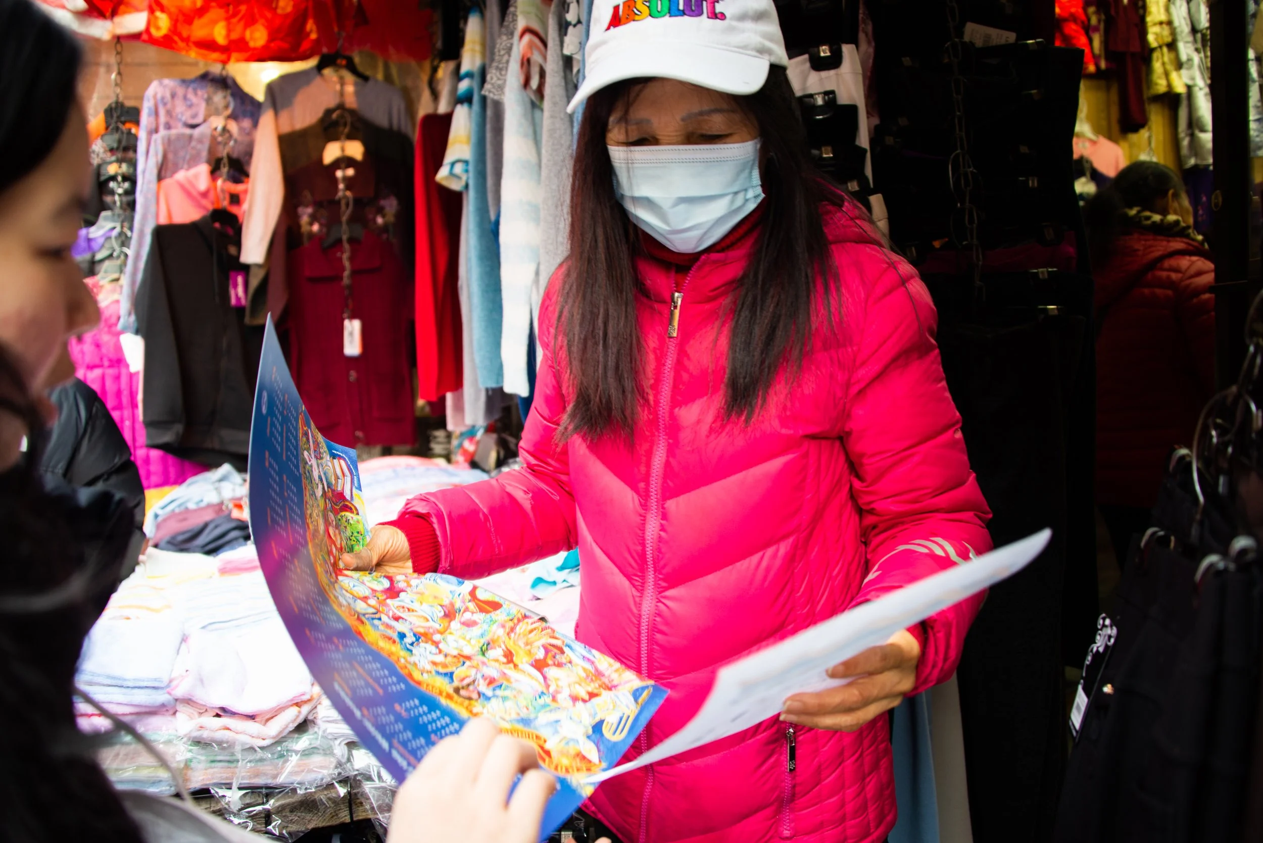Two women in a clothing store, one wearing a pink jacket and a white baseball cap with colorful text, the other an out-of-focus woman holding a brochure.
