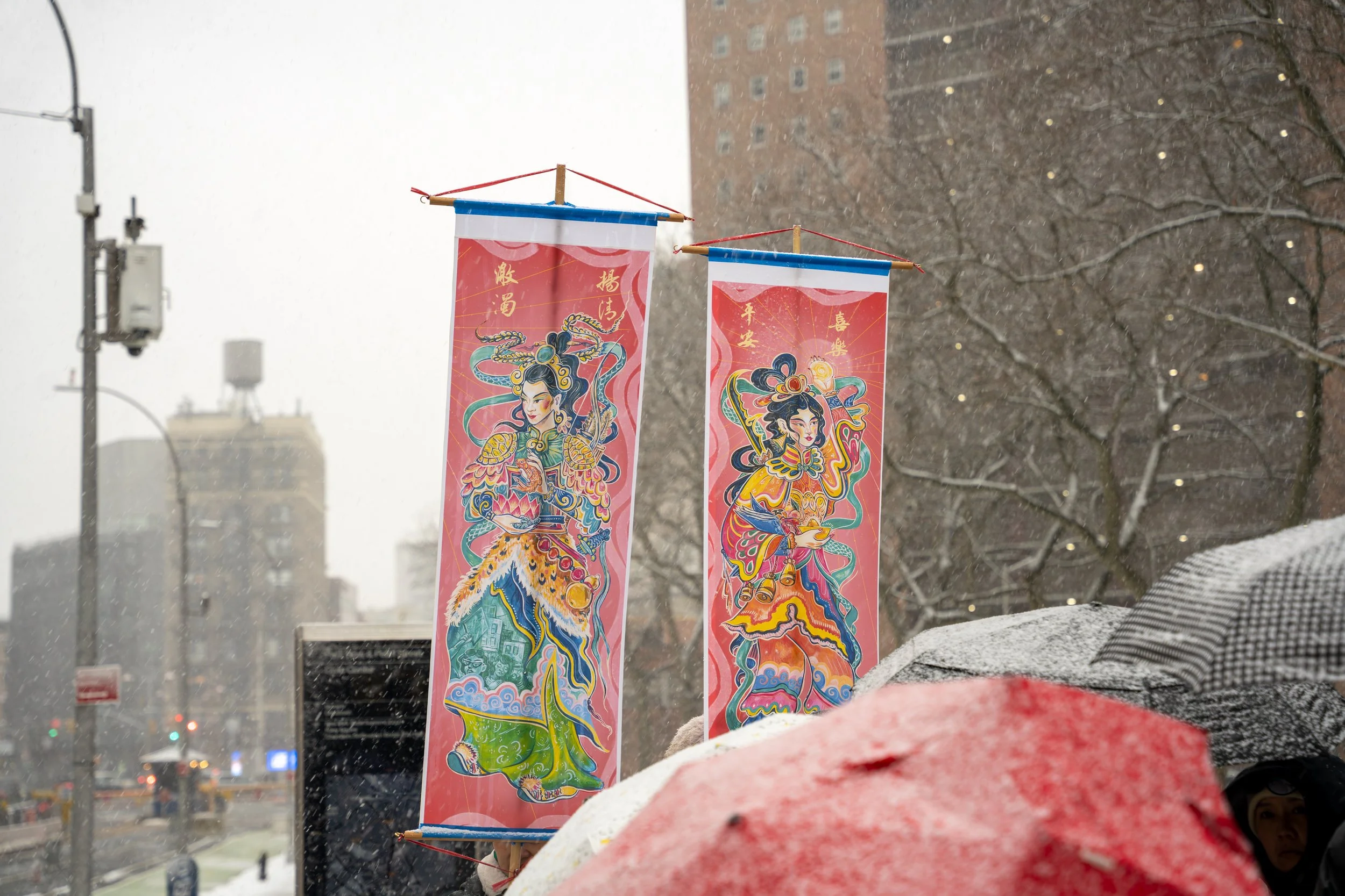 Two vertical banners featuring colorful traditional Chinese dragon and warrior artwork, held up outdoors in snowy weather, with people holding umbrellas and leafless trees in the background.