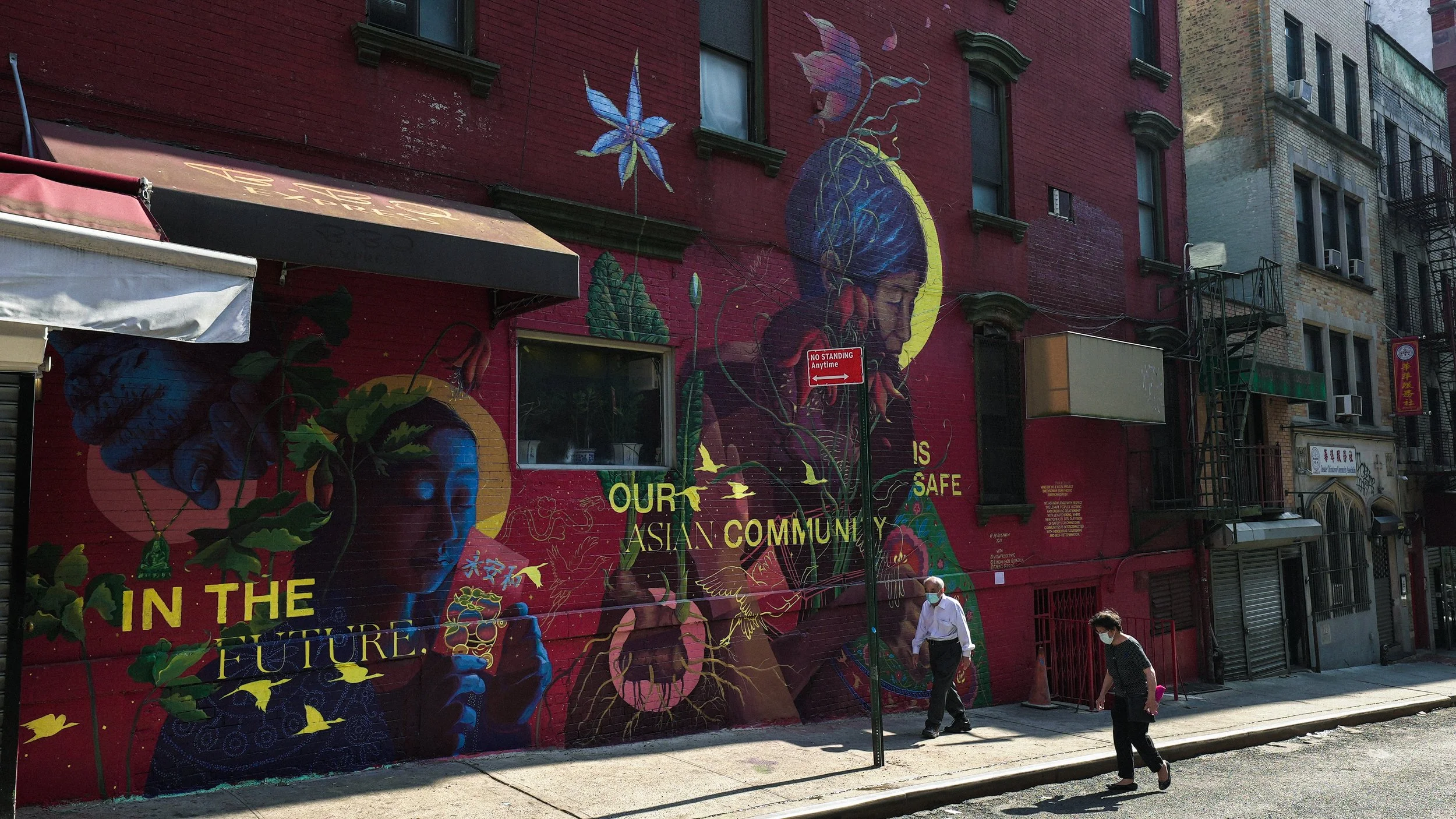 Colorful mural on a building featuring two Asian women with elaborate headpieces and traditional attire, surrounded by floral and abstract elements. The mural includes text that says 'In the Future Our Asian Community is Safe'. Two people wearing masks are walking past the mural on the sidewalk.
