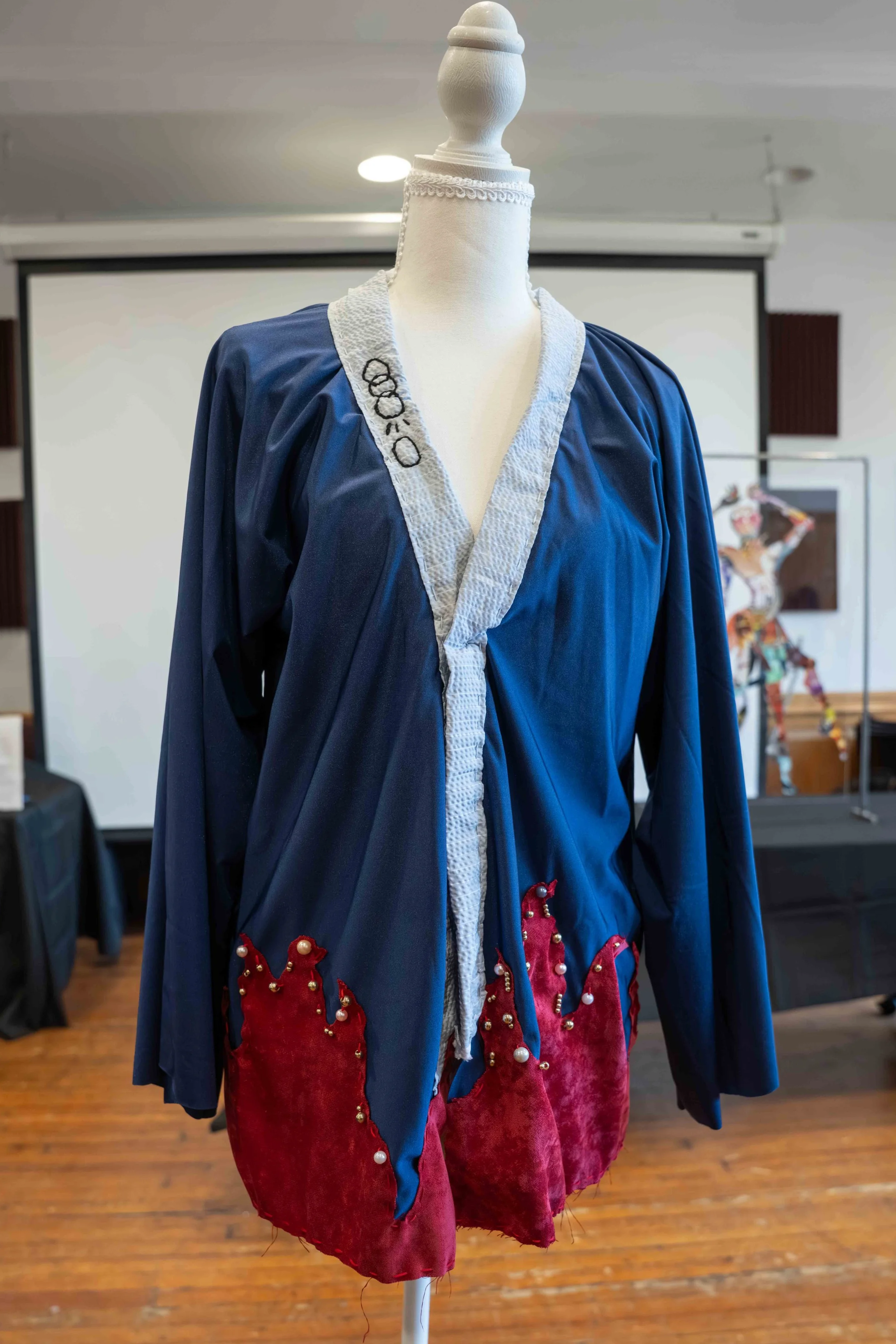 A mannequin dressed in a blue and red costume with decorative pearls and embroidery, displayed indoors with a blurred background of a room with wooden floors and artwork.