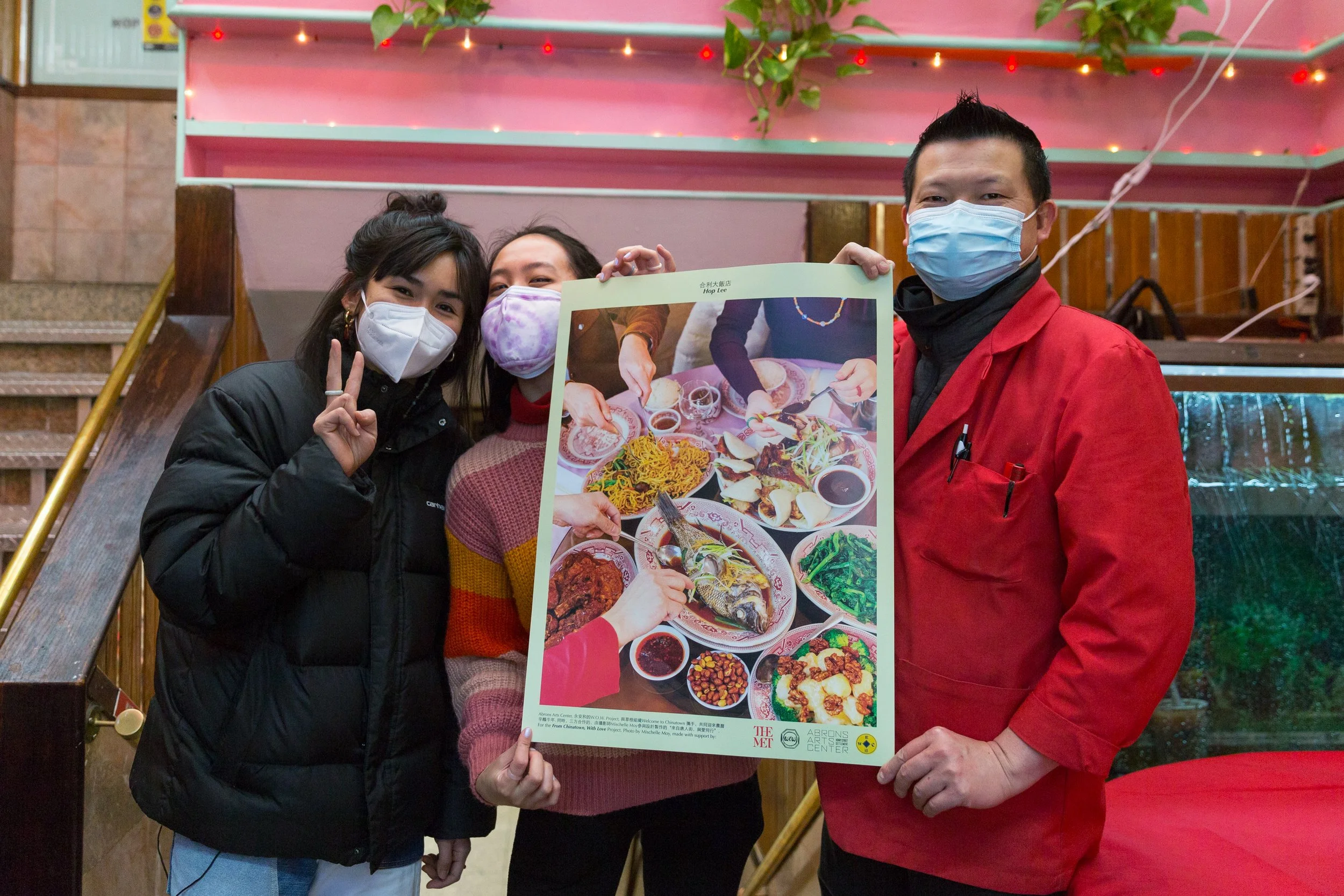 Three people wearing face masks, standing indoors with a pink wall and christmas lights. One person holds a poster showing various dishes including fish, noodles, and vegetables.