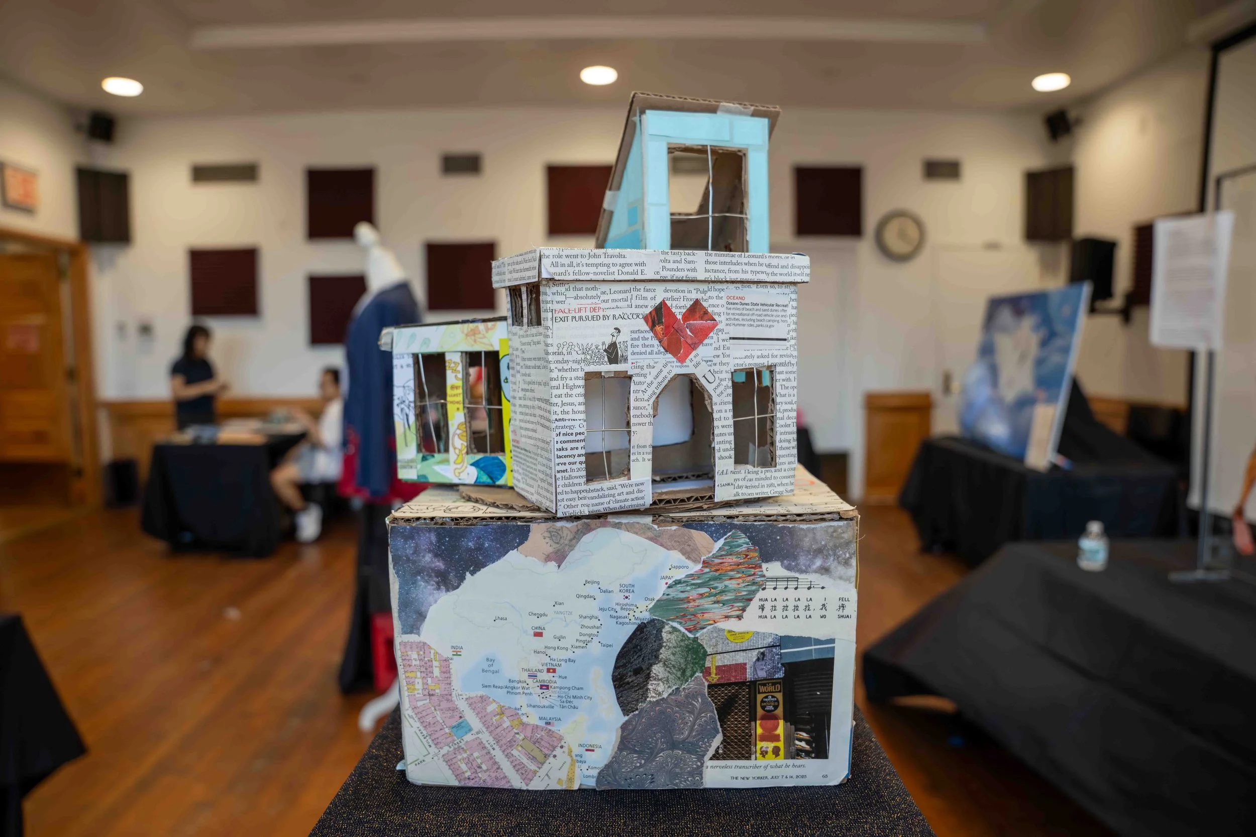 An intricate model made of cardboard and paper maps placed on a stand, featuring a small blue house-like structure on top, with a background of blurred conference or event room with people, tables, and display boards.