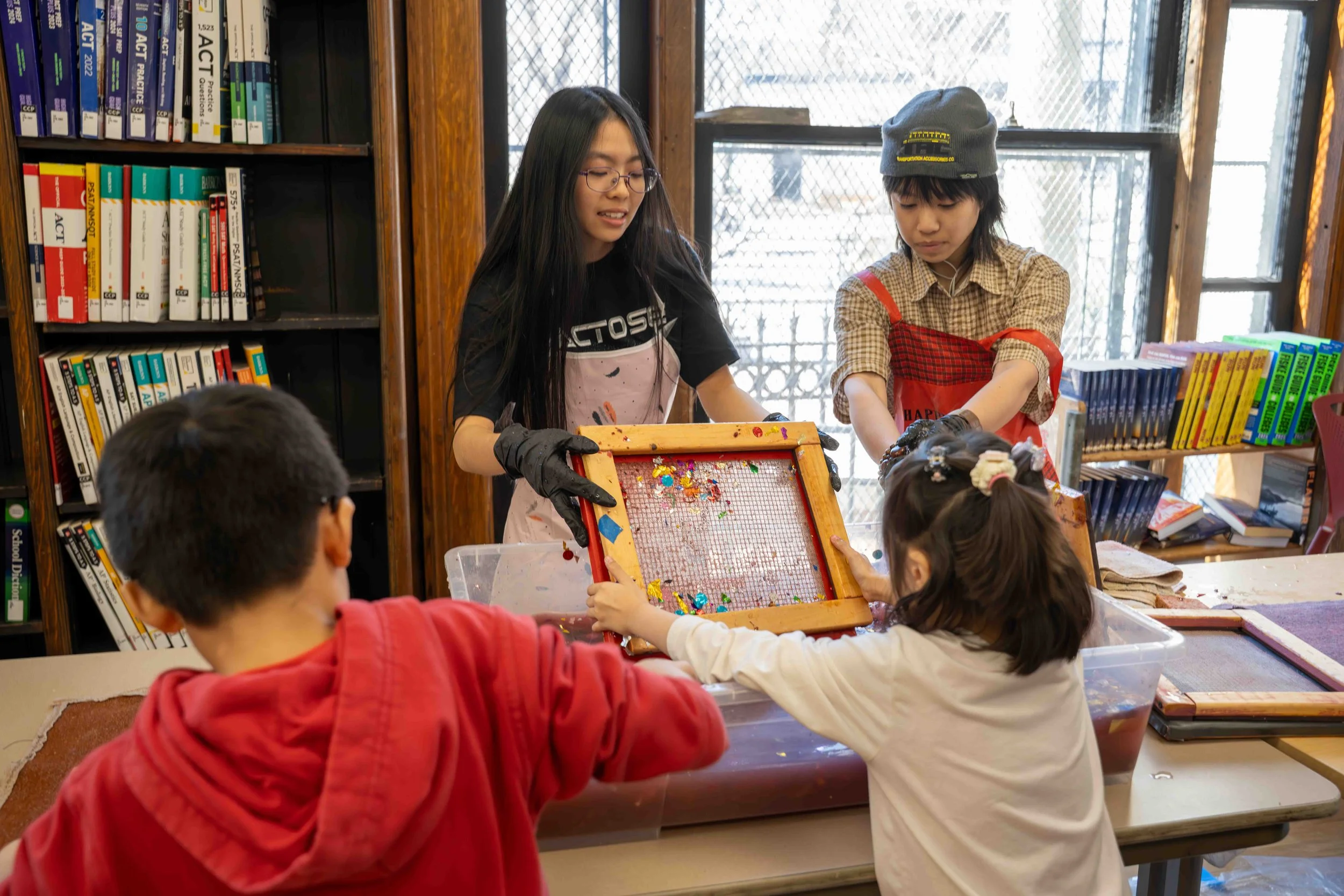 Two girls with gloves working at a craft table with two children, sorting colorful glitter and decorations in a classroom or library setting with bookshelves and windows.