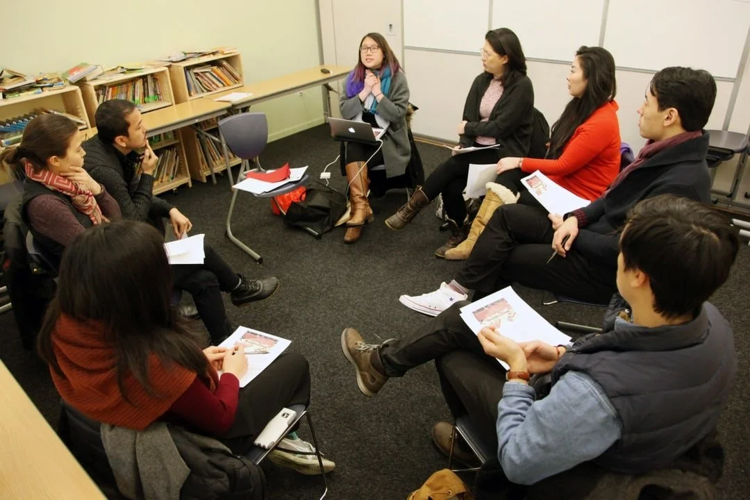 A group of eight people, mostly young adults, sitting in a circle in a small classroom or conference room, engaged in a discussion. Some are holding papers, and one woman is speaking.