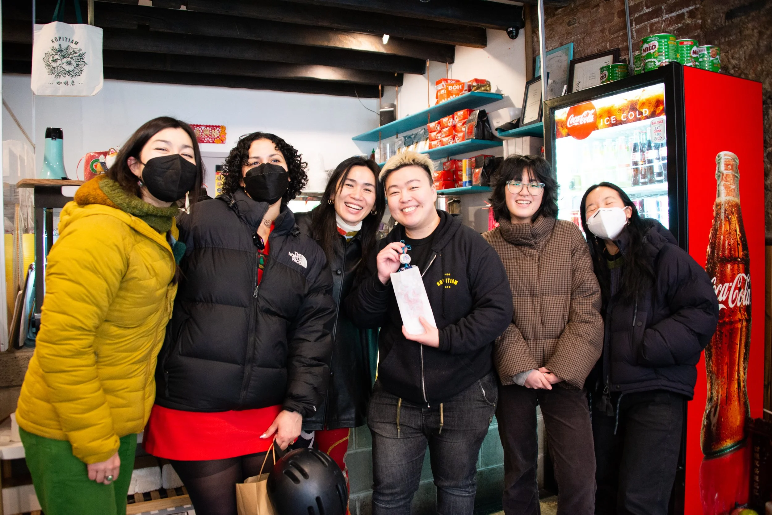 Group of six people wearing masks, standing in front of a refrigerator with drinks, smiling, inside a small shop or cafe.